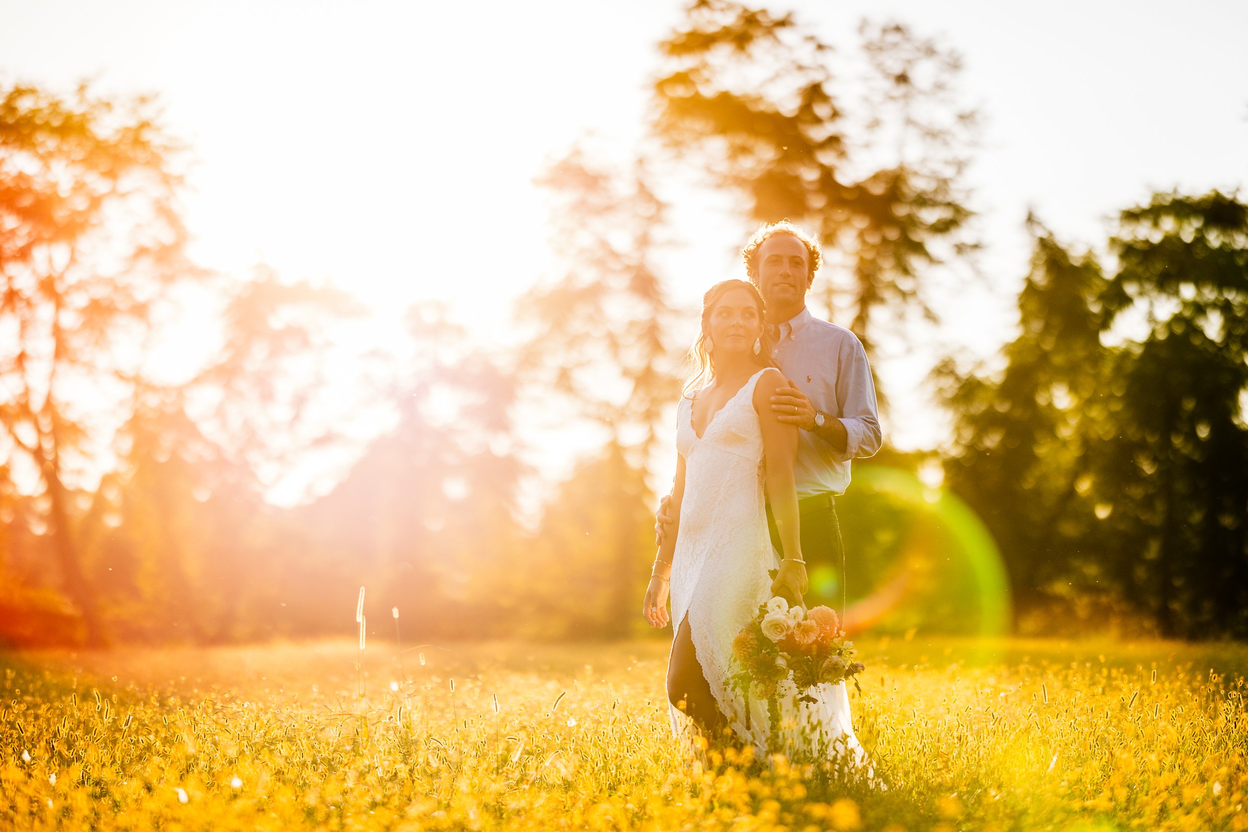A couple dressed in wedding attire standing in a sunlit field of yellow flowers, holding a bouquet, with trees and bright sunlight in the background.