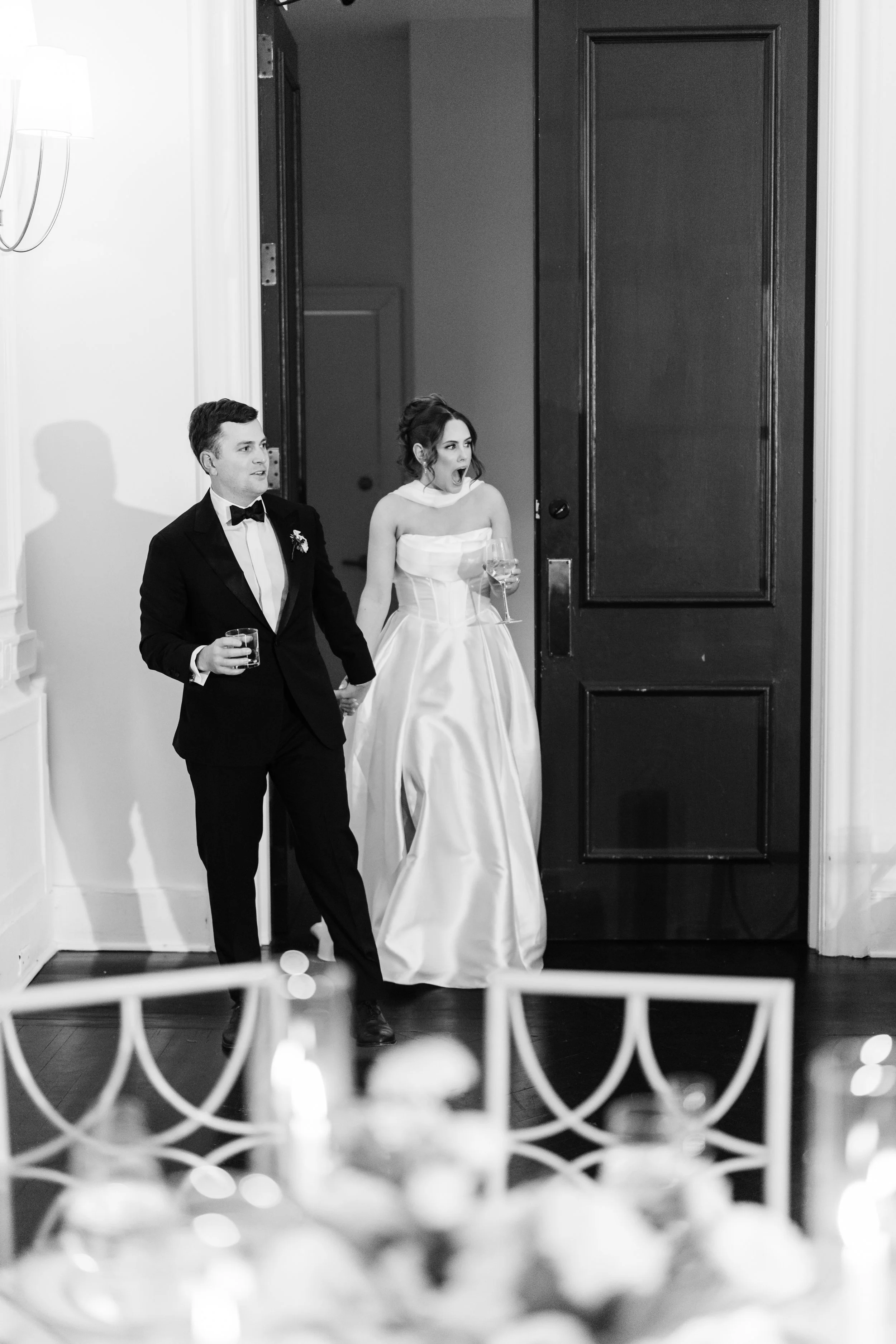 Black and white photo of a man and woman in formal wedding attire walking into a room, holding hands, with drinks in their hands.