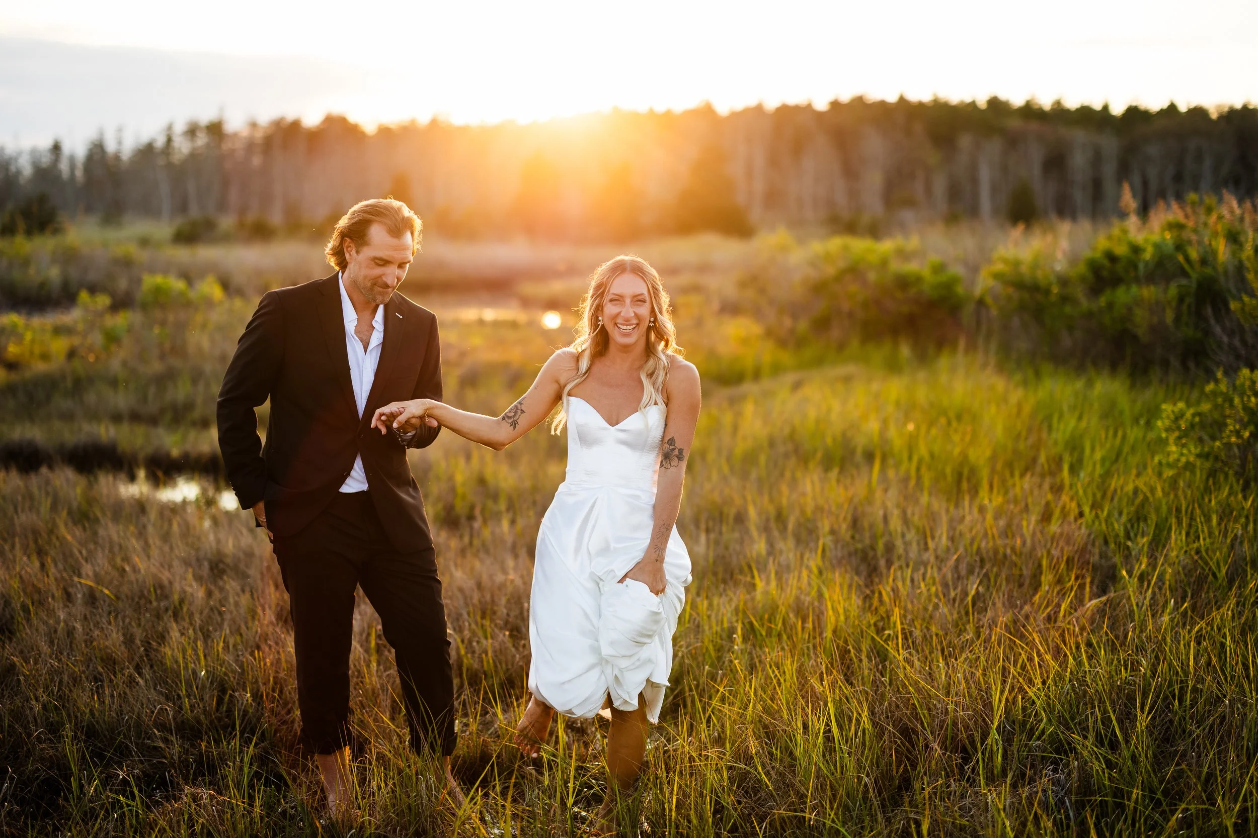 Man and woman walking through a grassy field at sunset, the woman is wearing a white dress and the man is in a black suit.