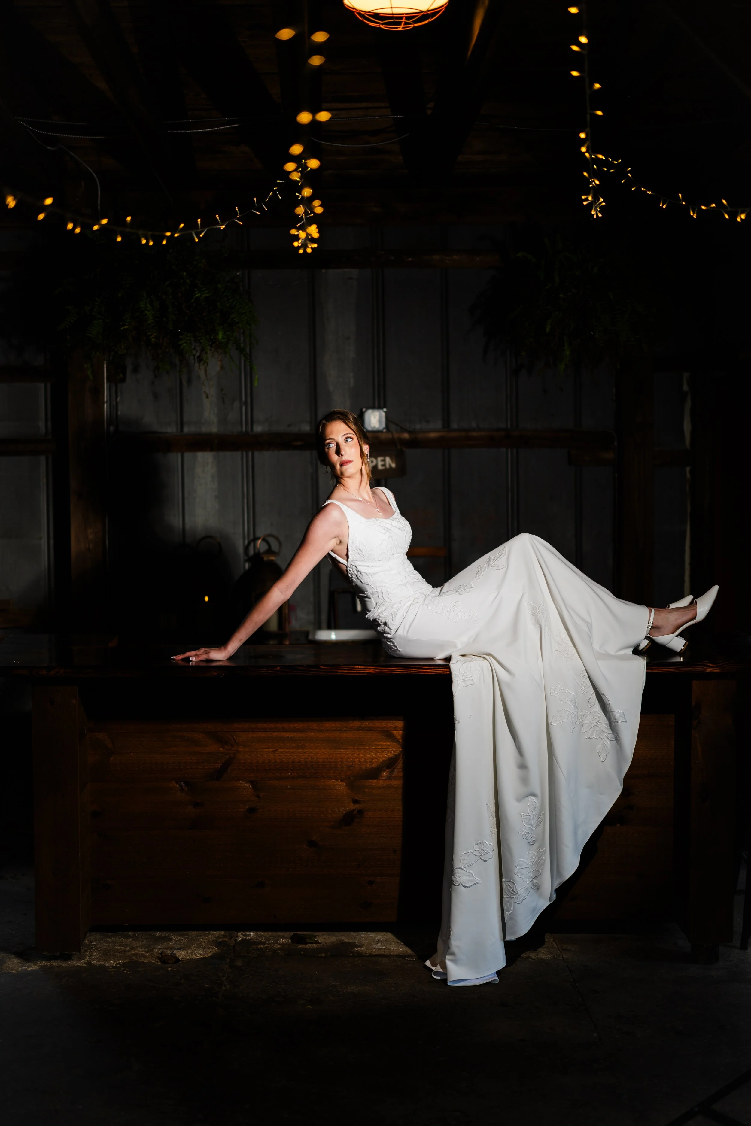 A woman in a white wedding dress and heels sitting on a wooden table in a dimly lit rustic setting with hanging string lights and greenery.