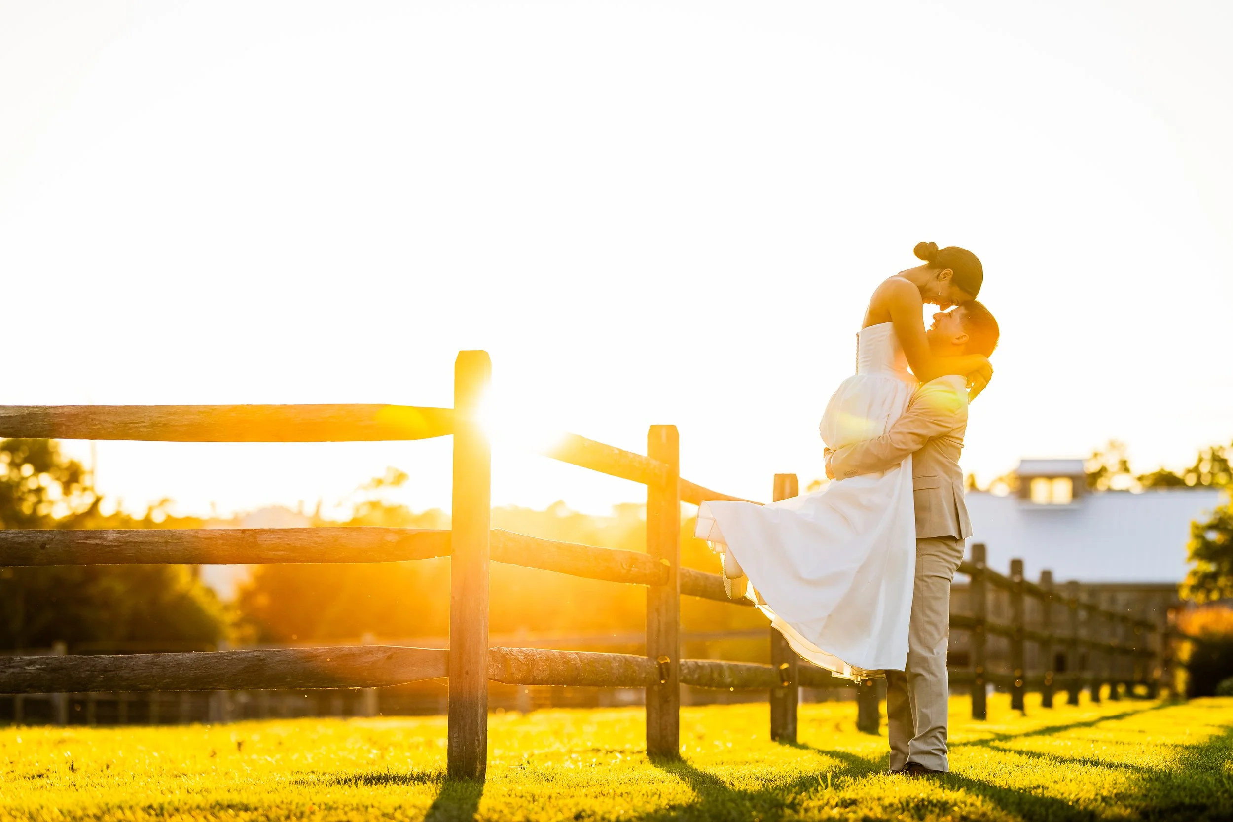 A couple at sunset, with the man lifting the woman in his arms, both dressed in wedding attire, in an outdoor setting with a wooden fence and green grass.