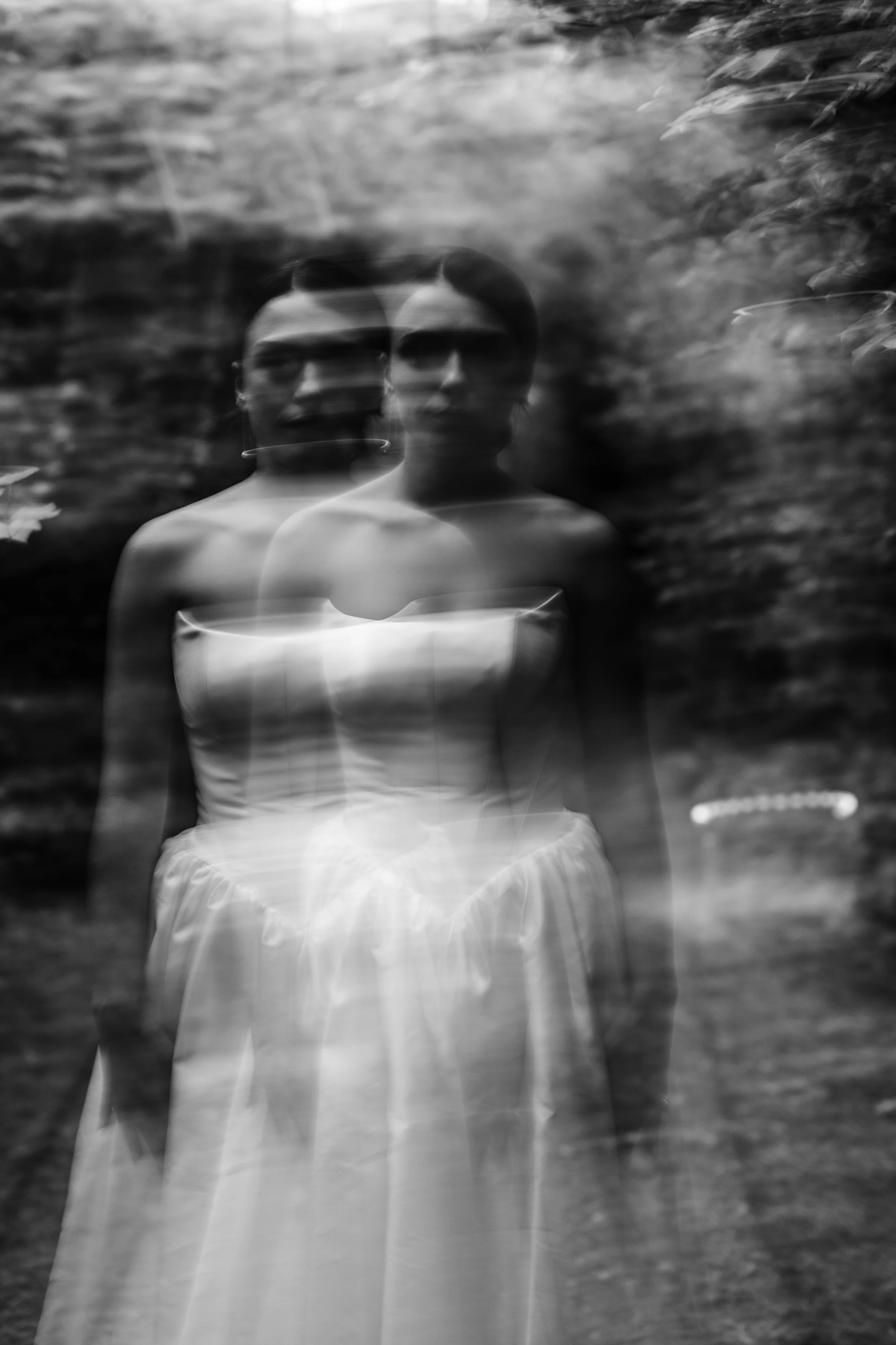 A double exposure black and white photograph of a woman standing outdoors, with blurred greenery in the background, and the woman's face appears as two overlapping images.