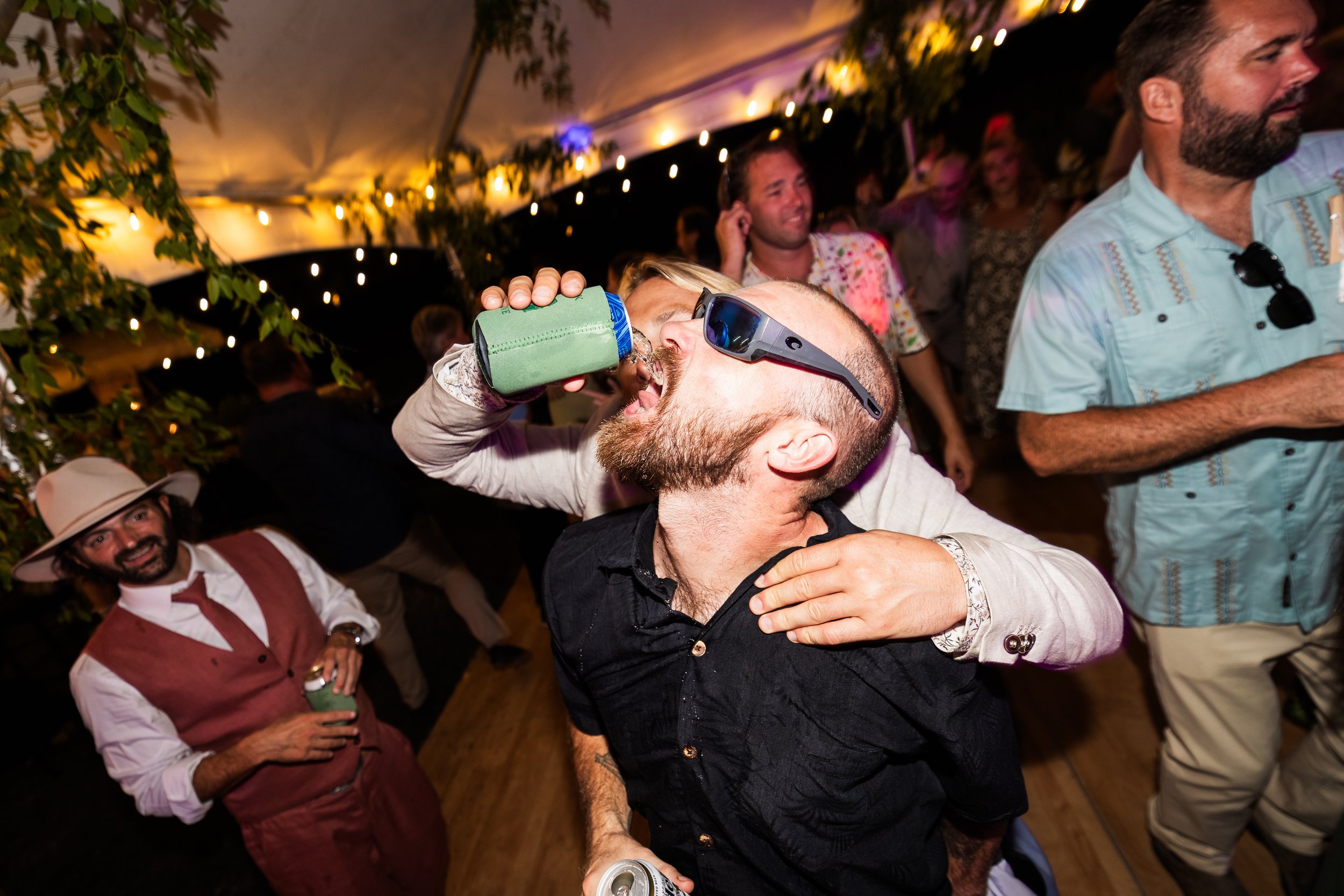 People partying at a lively event, with one person drinking from a green bottle while wearing sunglasses, and others smiling and holding drinks under string lights in a decorated tent.