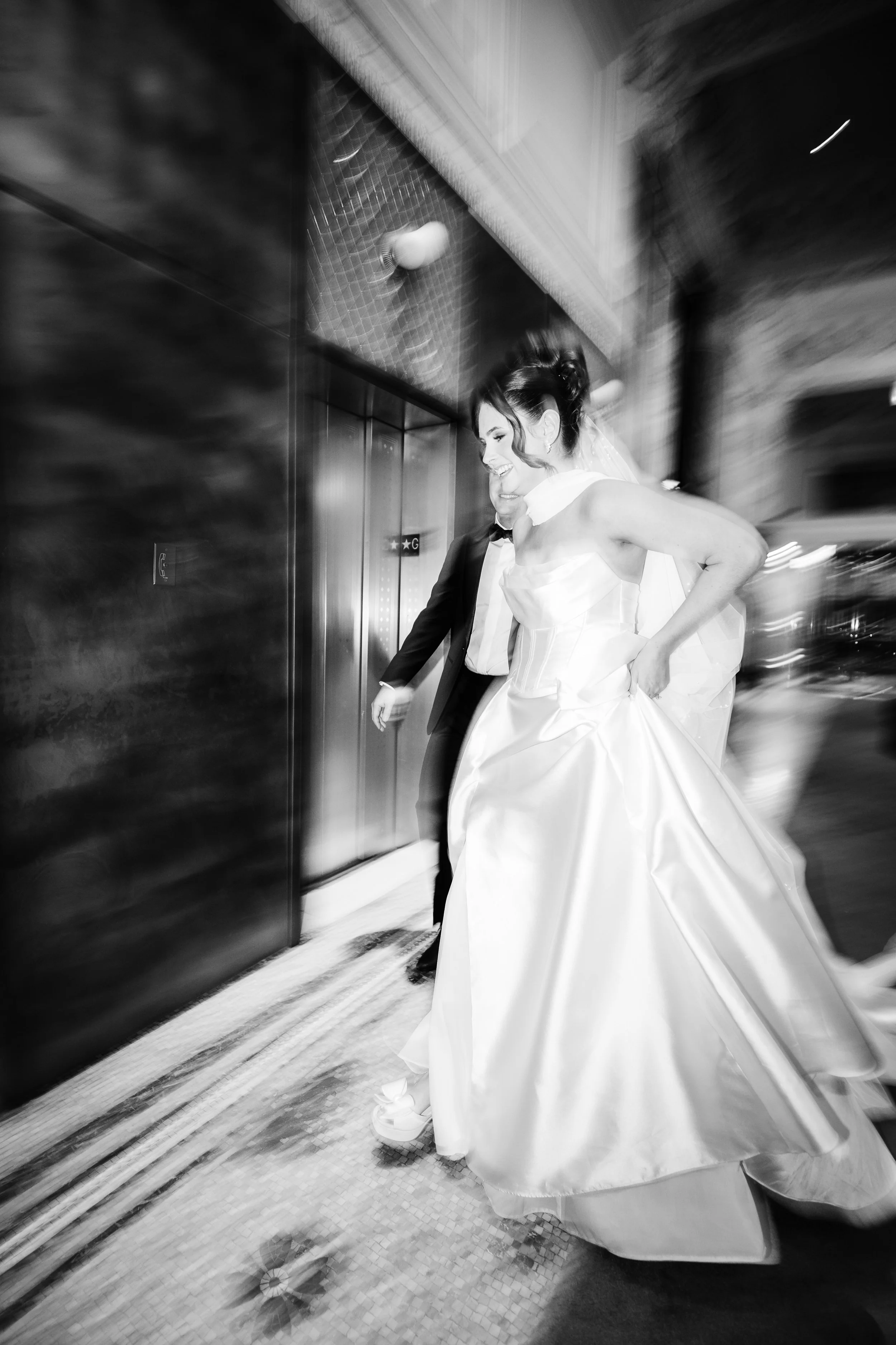 Black and white photo of a smiling bride in a wedding gown and veil walking outside with a groom in a suit and bowtie.
