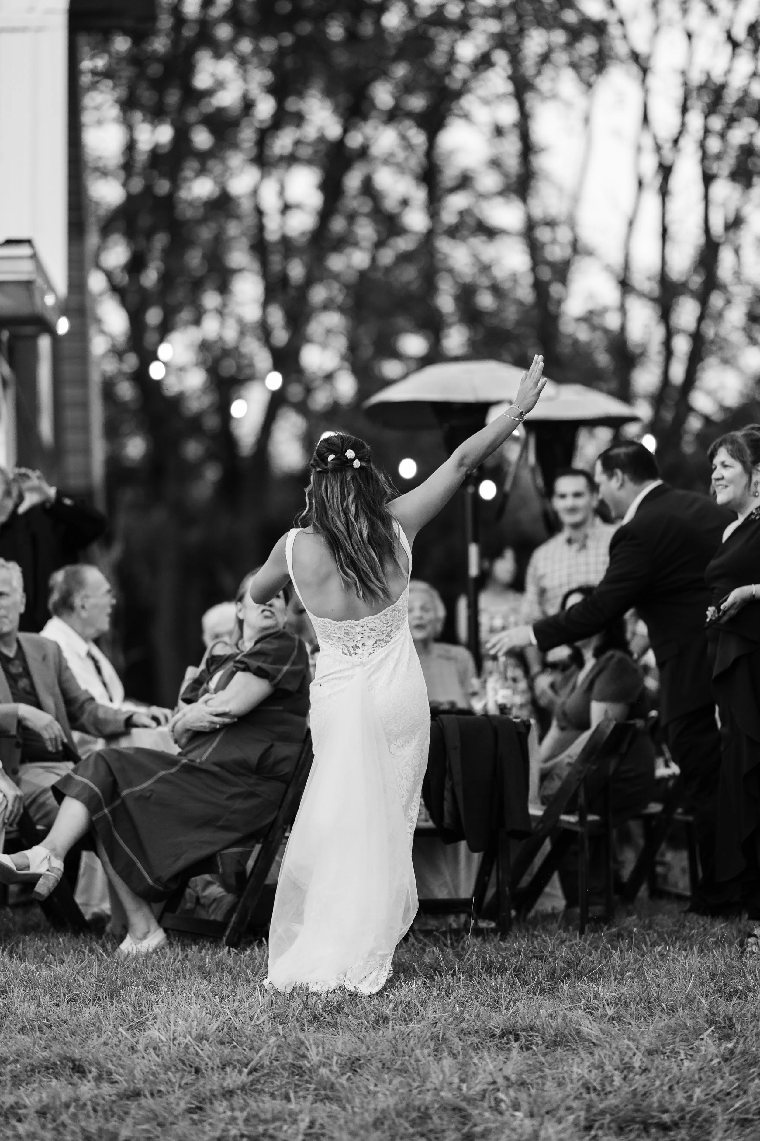 A woman in a wedding dress dancing at an outdoor wedding reception with guests seated around her, some clapping and enjoying the moment, with trees in the background and evening lights.