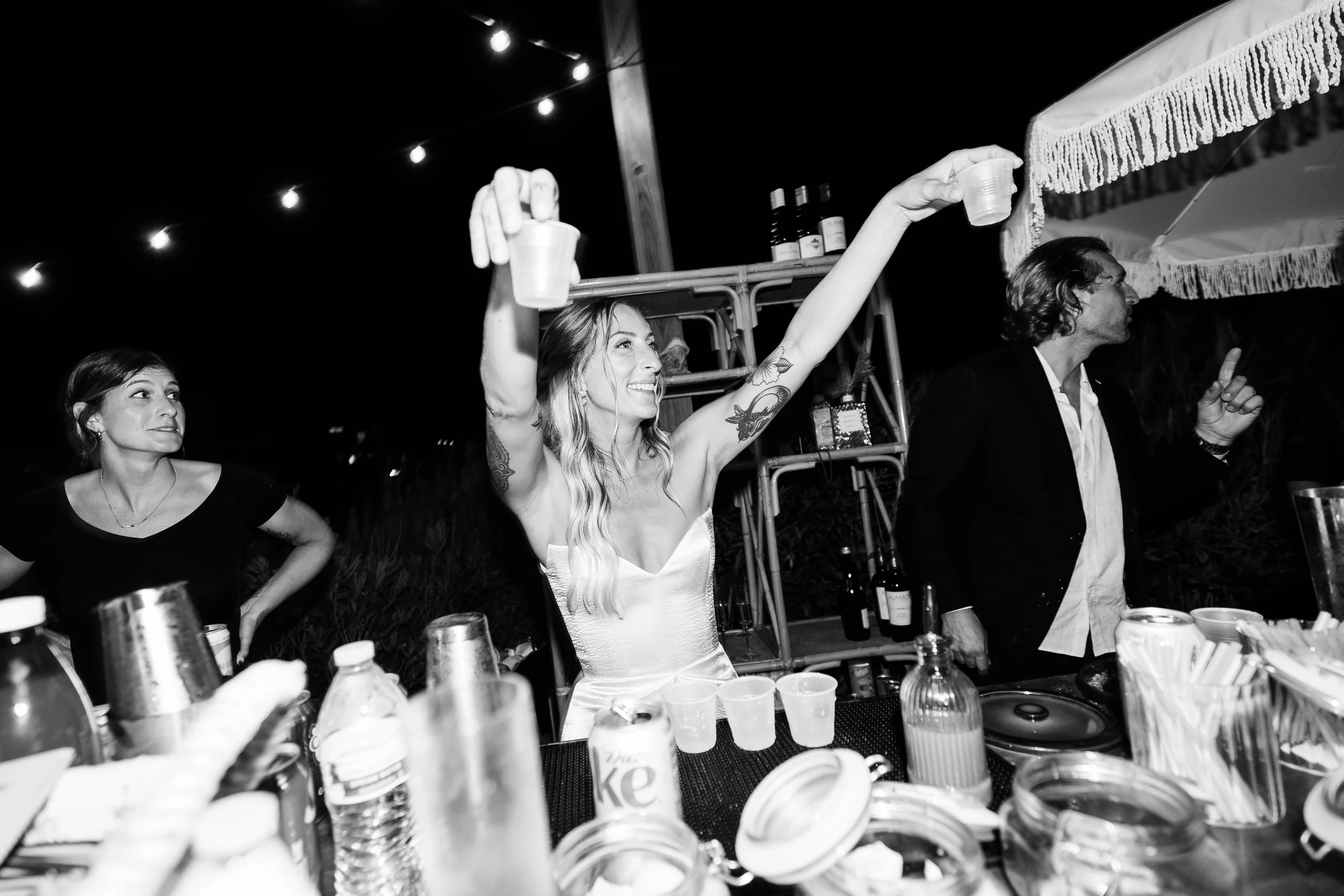A woman in a white dress smiling and throwing her arms in the air at a party, with two other people nearby, surrounded by drinks and bar supplies under string lights at night.