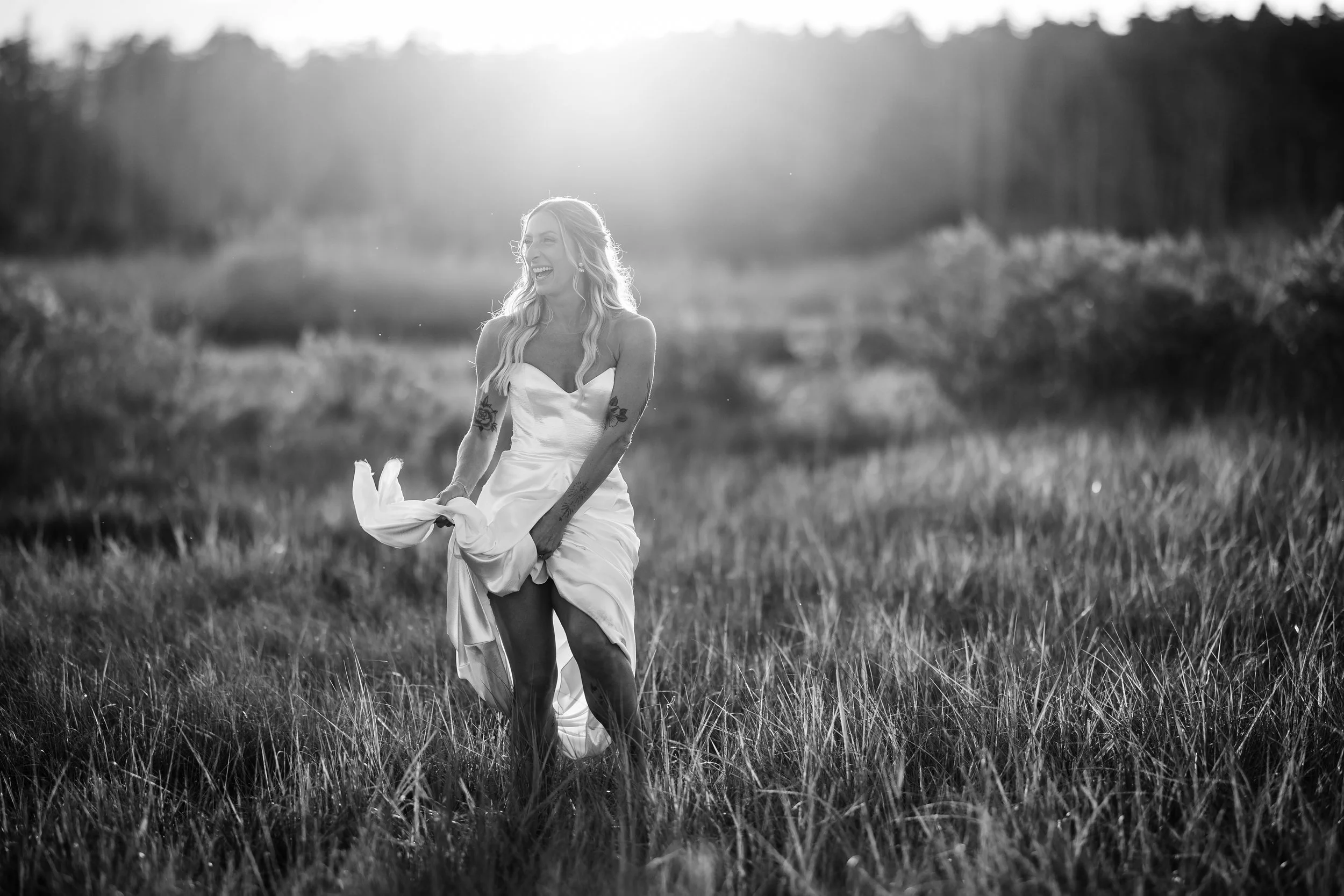 A woman in a wedding dress laughing and holding up her dress in a field with tall grass, sunlight shining in the background.