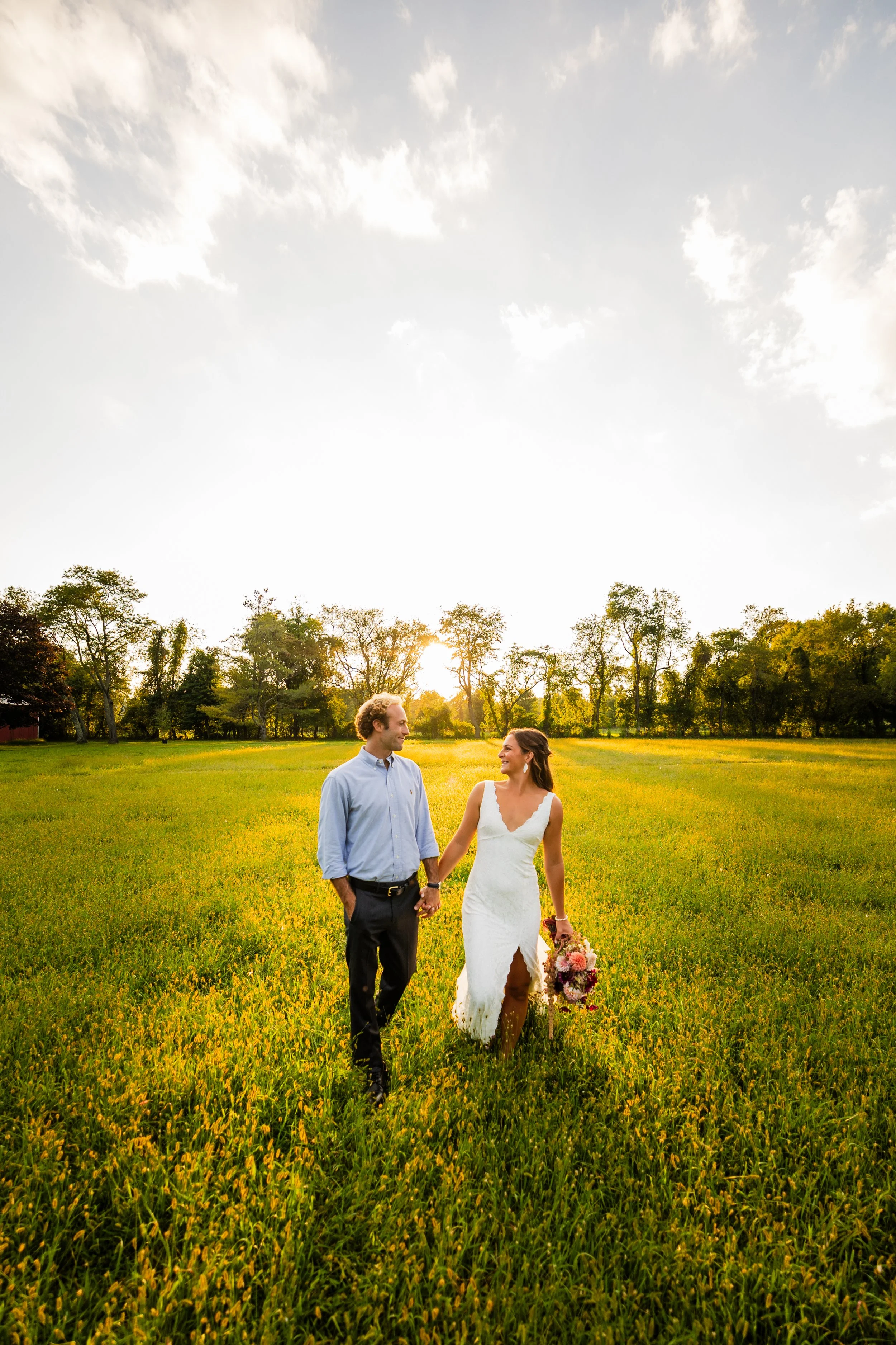 A couple walking hand in hand through a green field during sunset, with trees and a partly cloudy sky in the background.