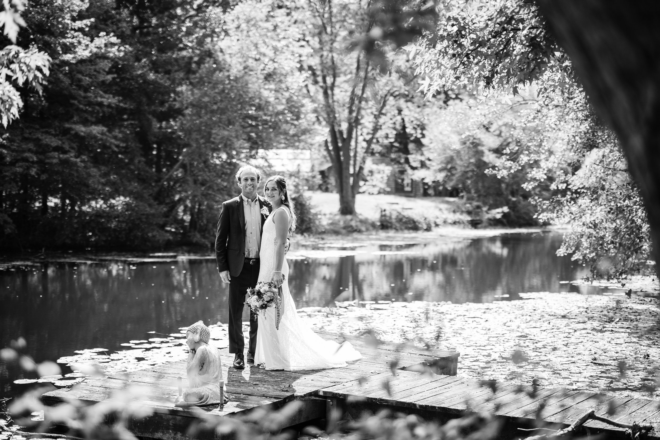A bride and groom standing on a wooden dock beside a pond with lily pads, surrounded by trees in a park during daytime, in black and white.