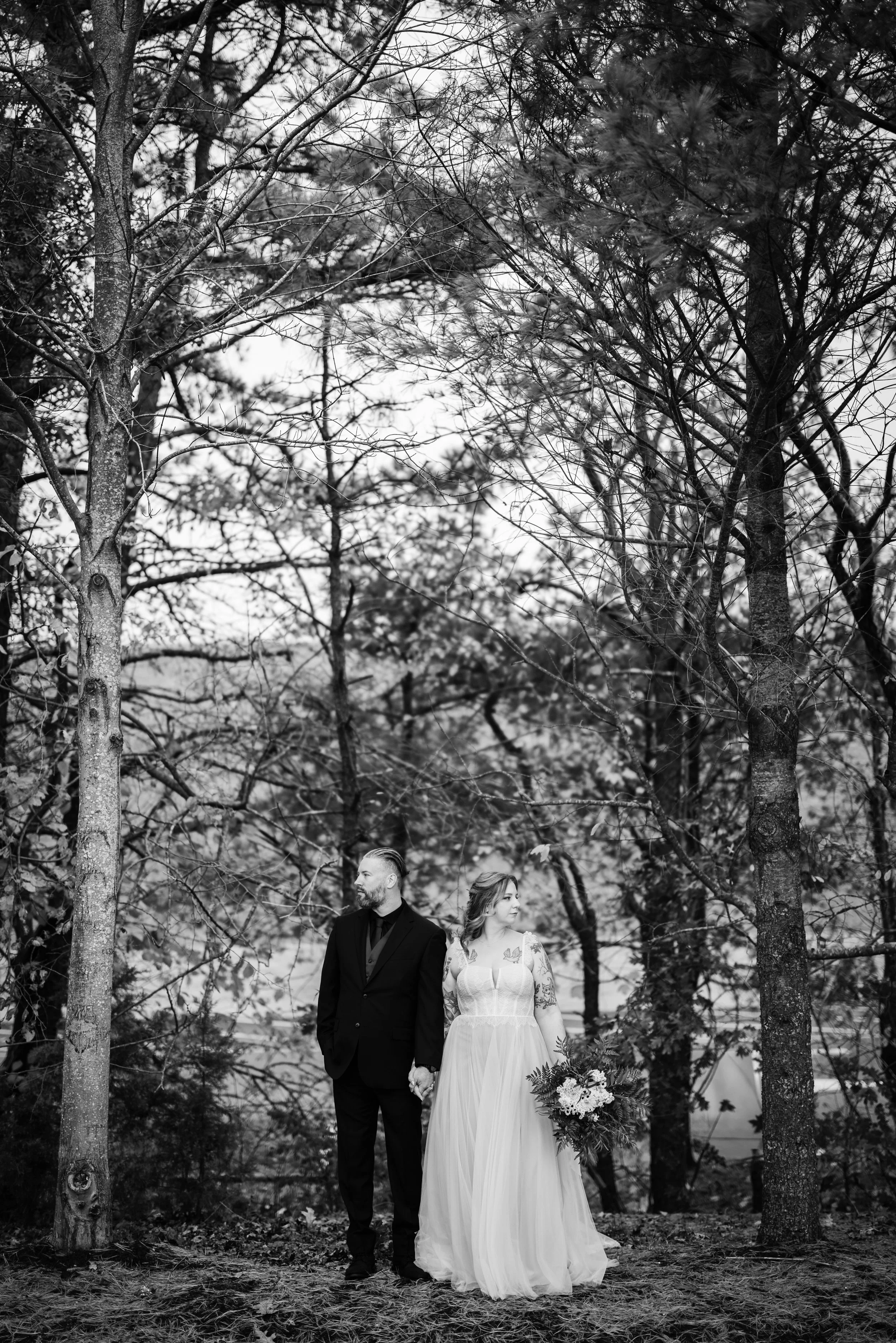 A bride and groom holding hands and standing among trees in a forest, with the bride holding a bouquet, captured in black and white.