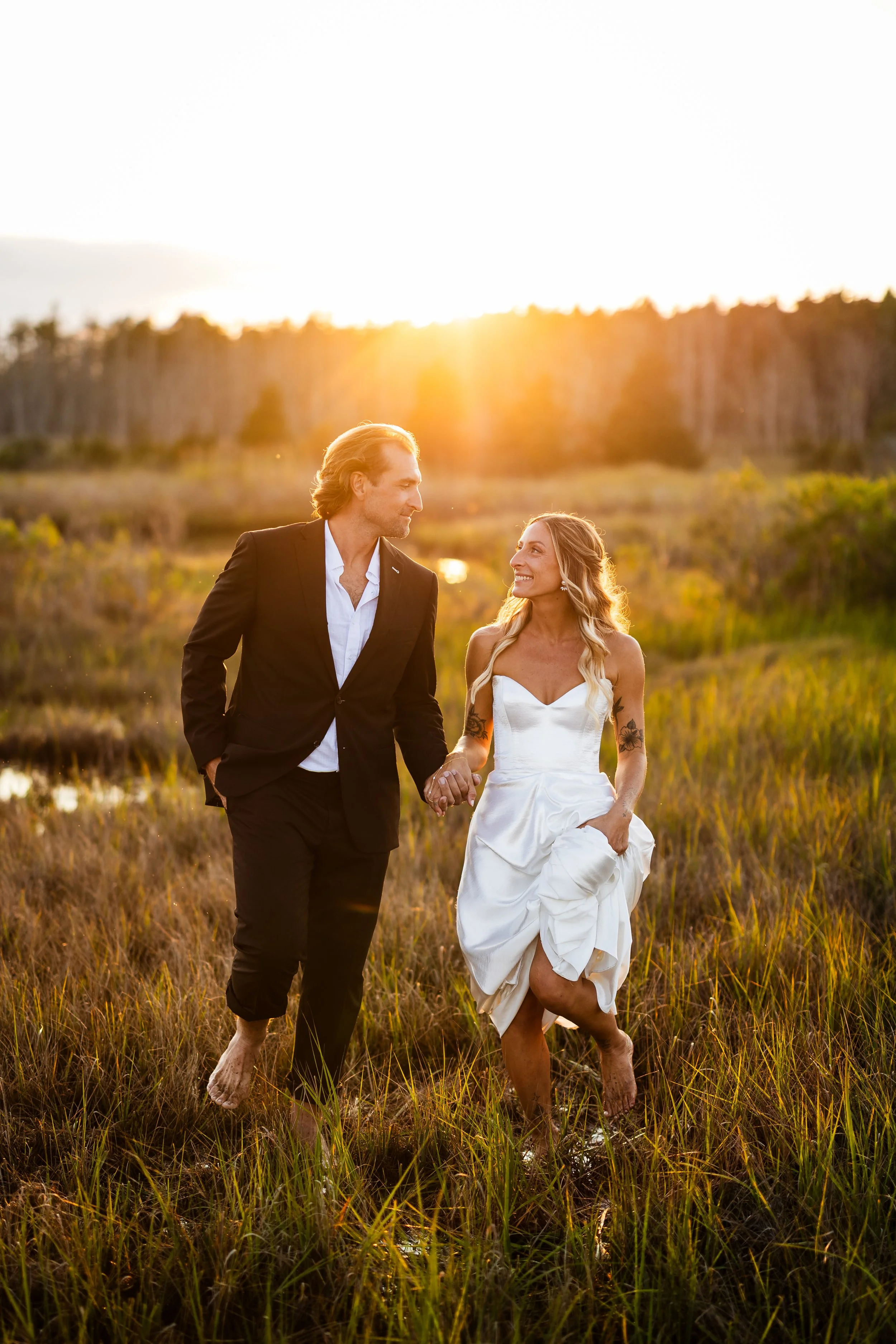 A couple walking hand in hand through a marsh at sunset, with the woman in a white dress and the man in a black suit, both barefoot and smiling.