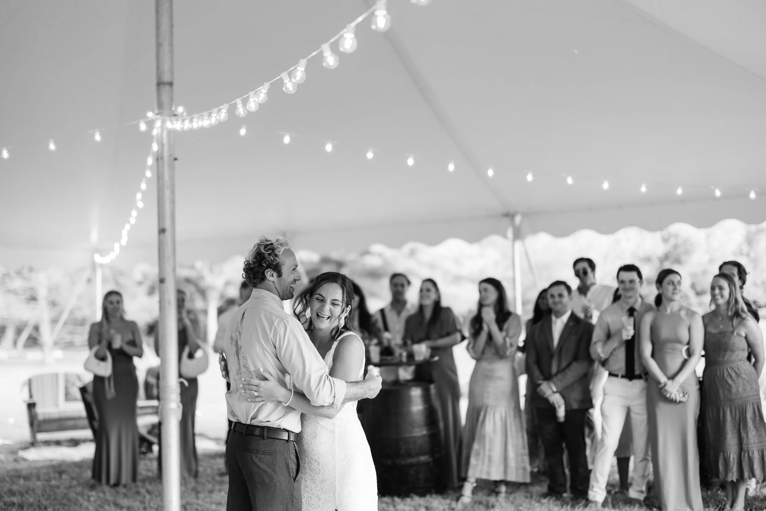 Couple dancing at a wedding reception under a tent with string lights, while guests watch and smile in the background.