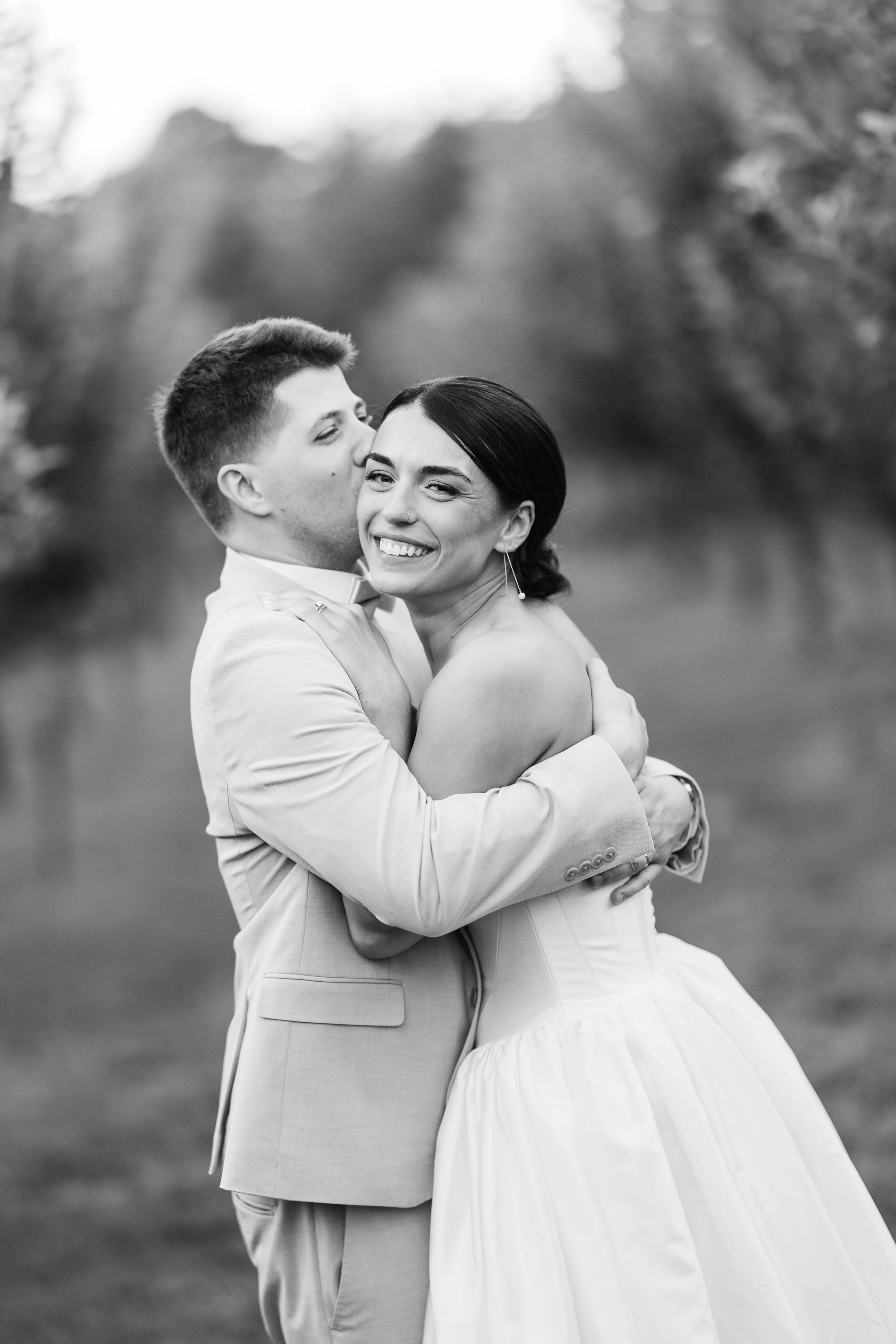 A black and white photo of a bride and groom embracing outdoors, with the groom kissing the bride on the cheek and the bride smiling.