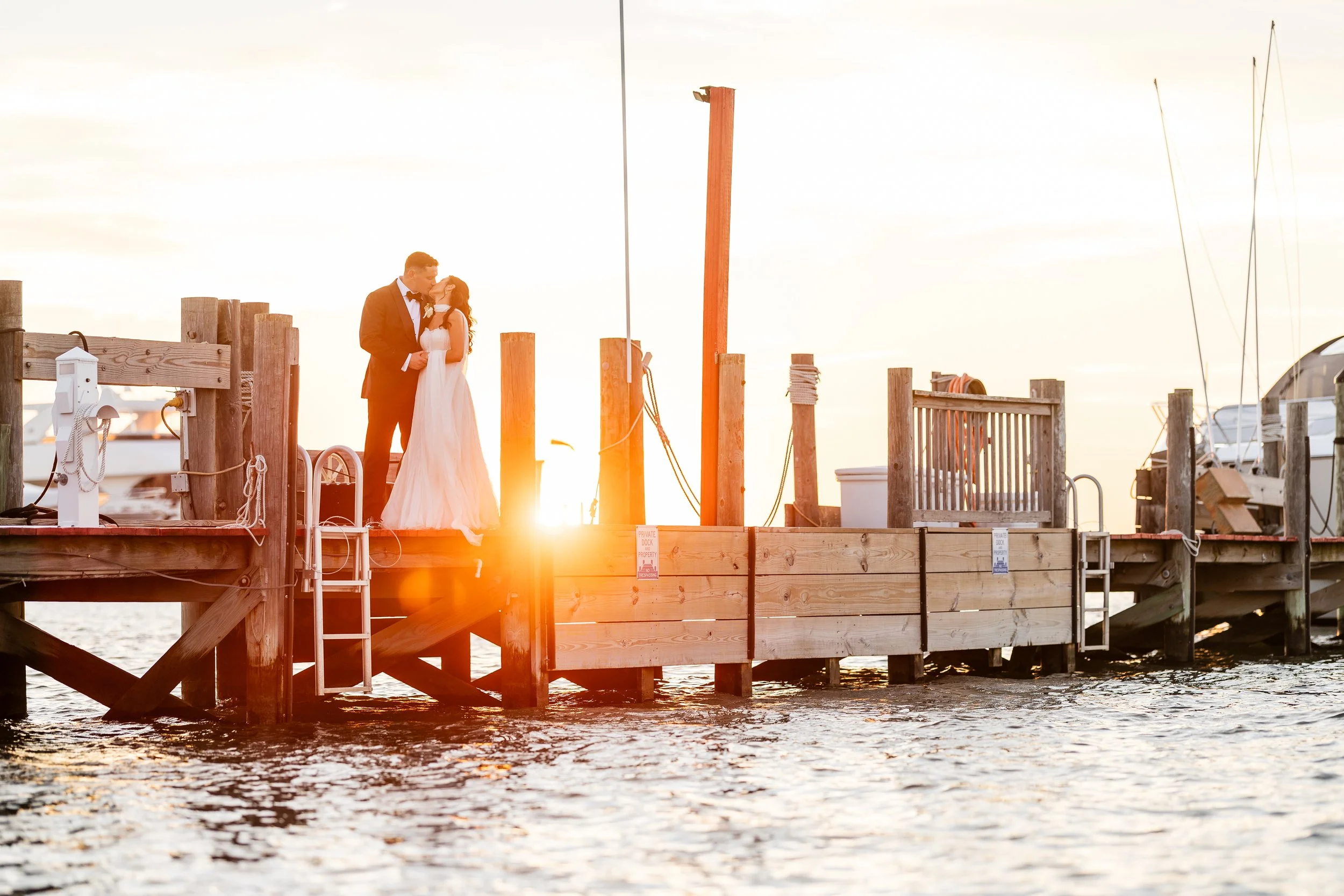 Bride and groom sharing a kiss on a wooden dock at sunset, with boats and water in the background.