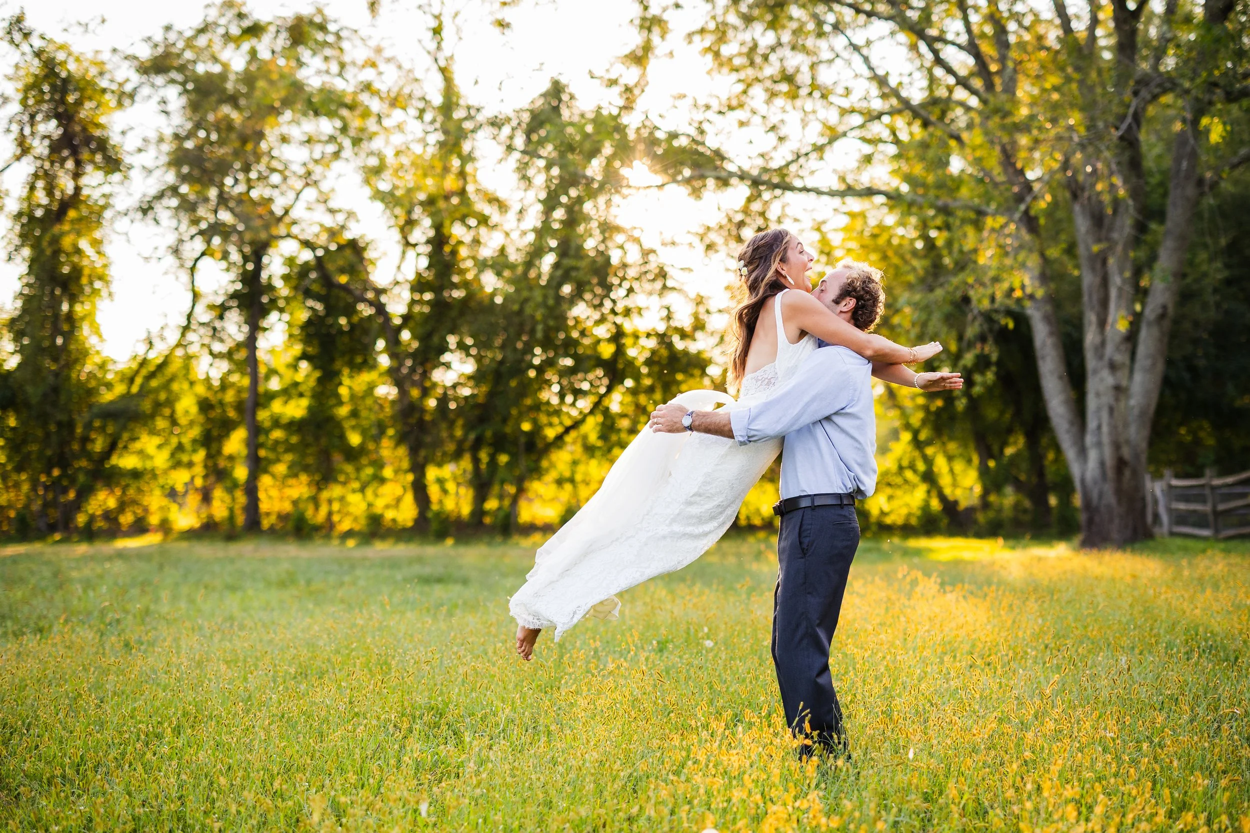 A man lifting a woman in a white dress in a grassy field during sunset with trees in the background.