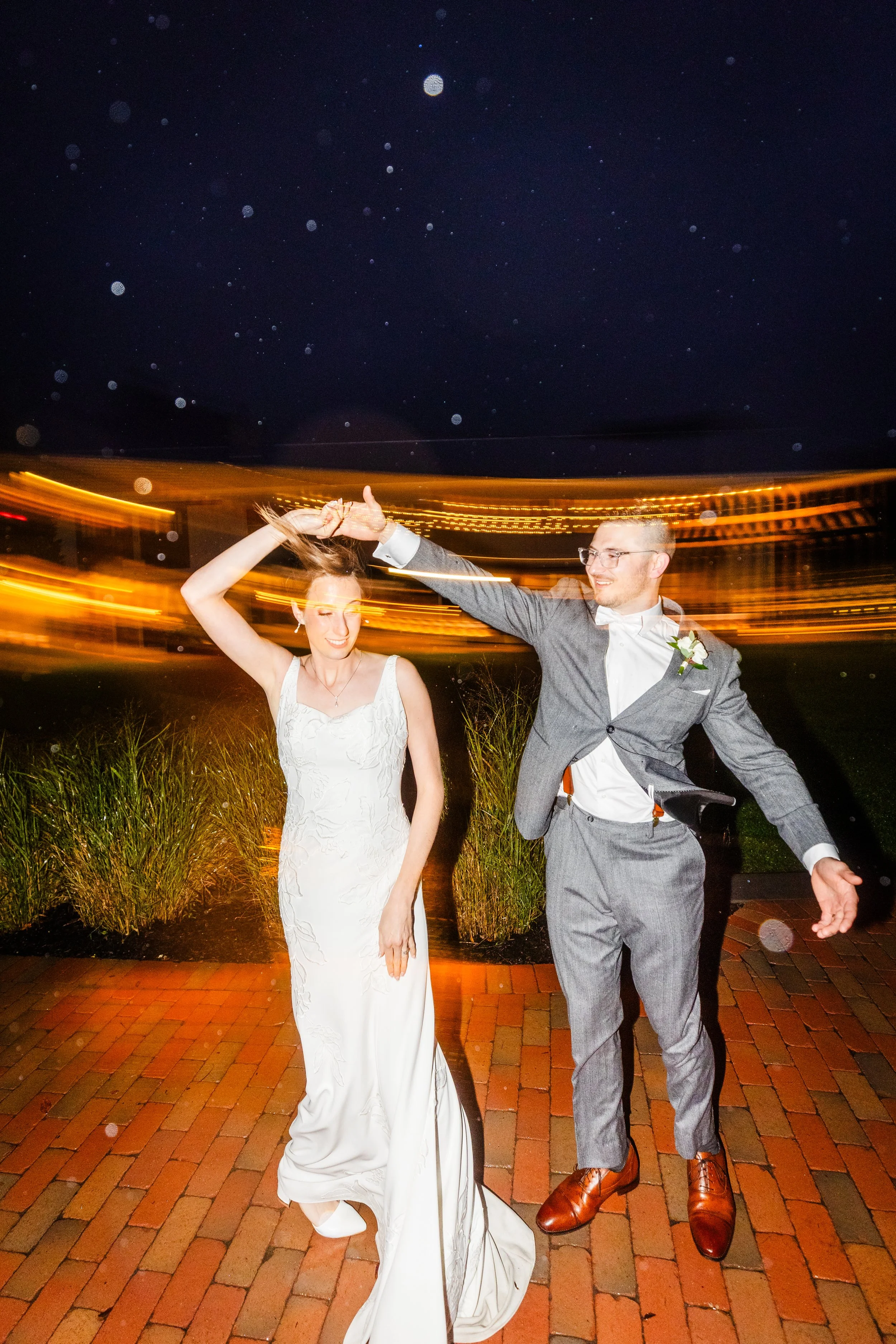 A bride and groom dancing, holding hands, during their wedding celebration at night with blurred lights in the background.