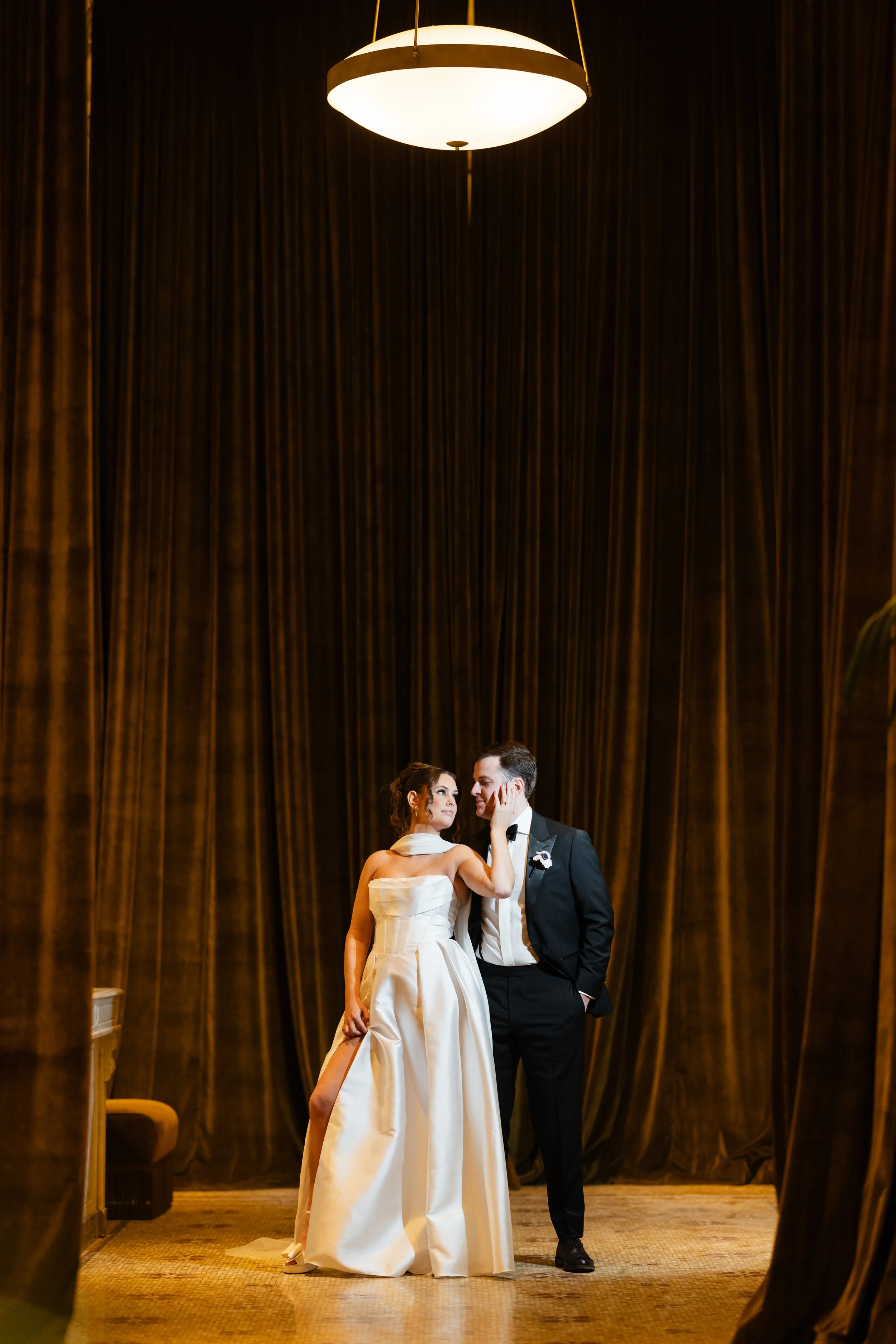 A couple dressed in formal wedding attire standing close together in front of a dark curtain with warm lighting overhead. The woman is touching the man's face and wearing a white gown, while the man is in a black tuxedo with a white shirt and bow tie