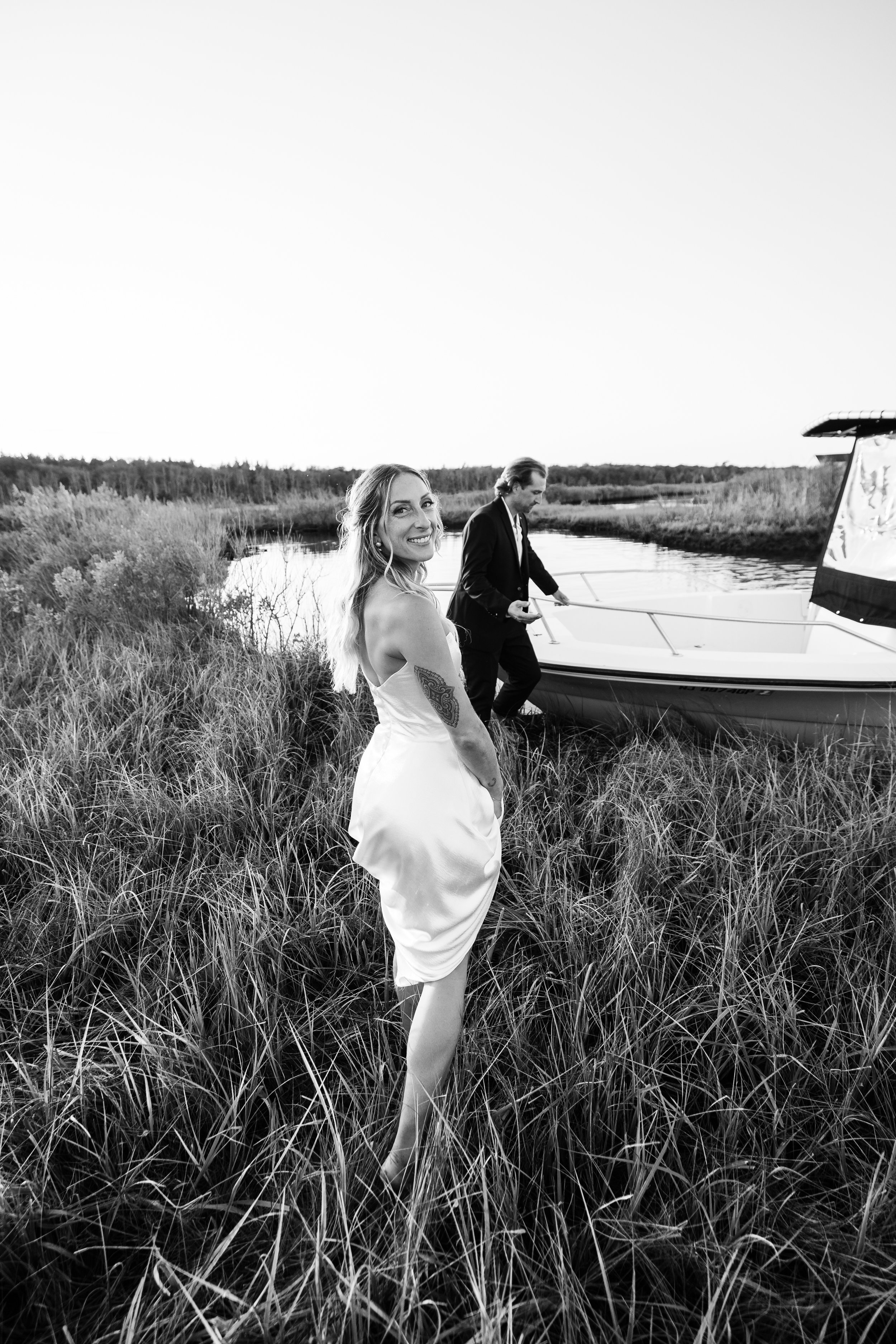 A woman in a white dress standing in tall grass near a boat, with a man in a suit preparing the boat, set in a natural landscape with water and trees in the background, black and white photograph.