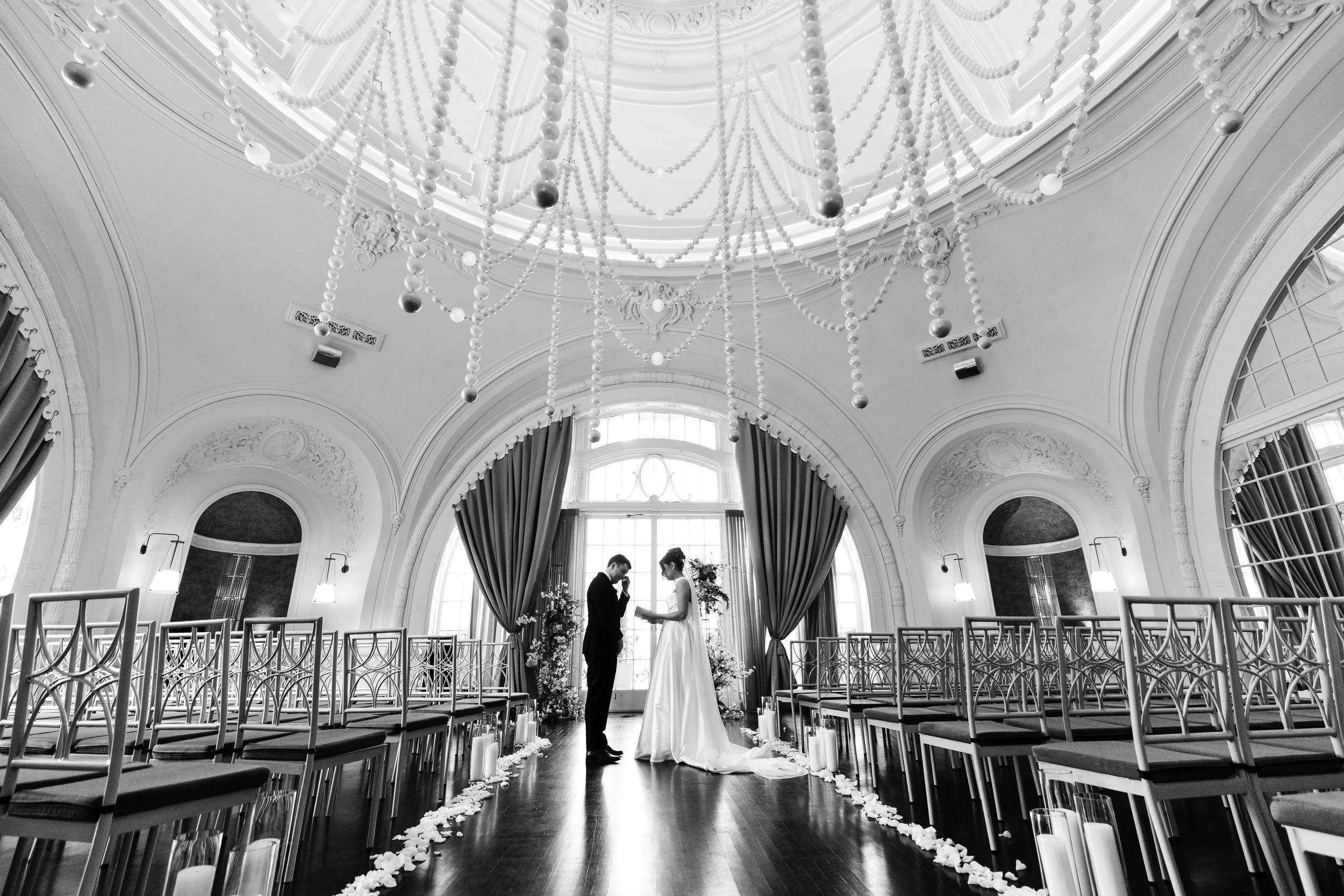 A black and white photo of a wedding ceremony inside an elegant, ornate hall with high ceilings and large windows. A couple is standing in front of the window, holding hands, with the bride in a long dress and the groom in a suit. The aisle is decora