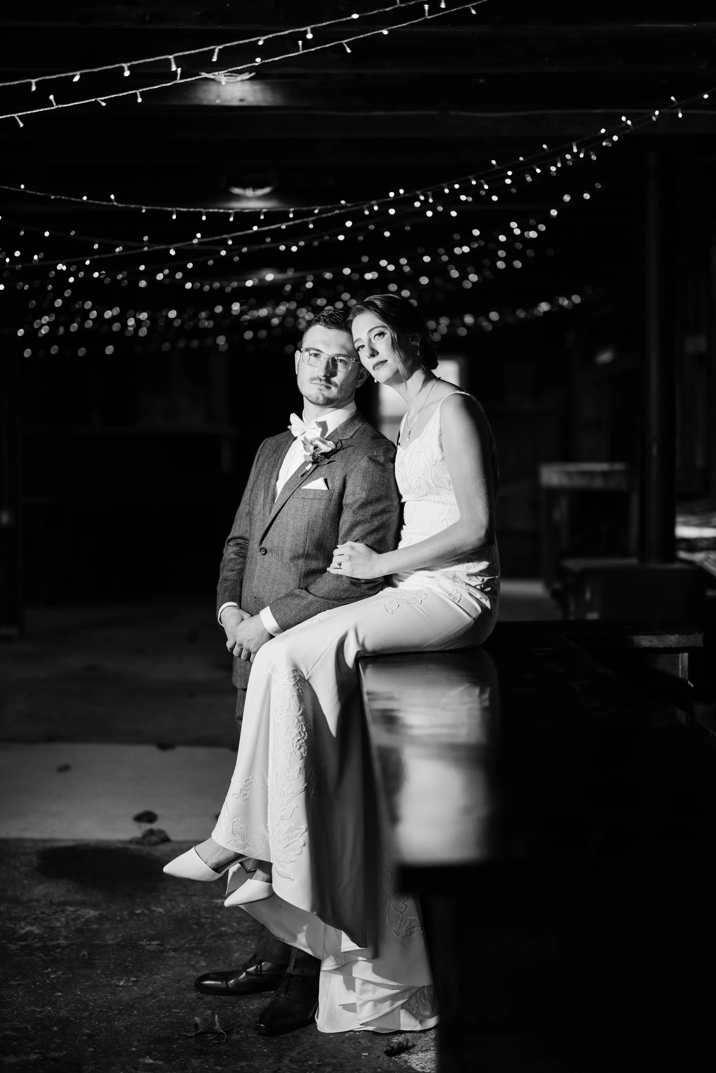 Black and white photo of a couple at a wedding, with the woman seated on a bench or ledge and the man standing beside her. The woman wears a white wedding dress and the man a suit with a boutonniere. They are in a dimly lit space with string lights h