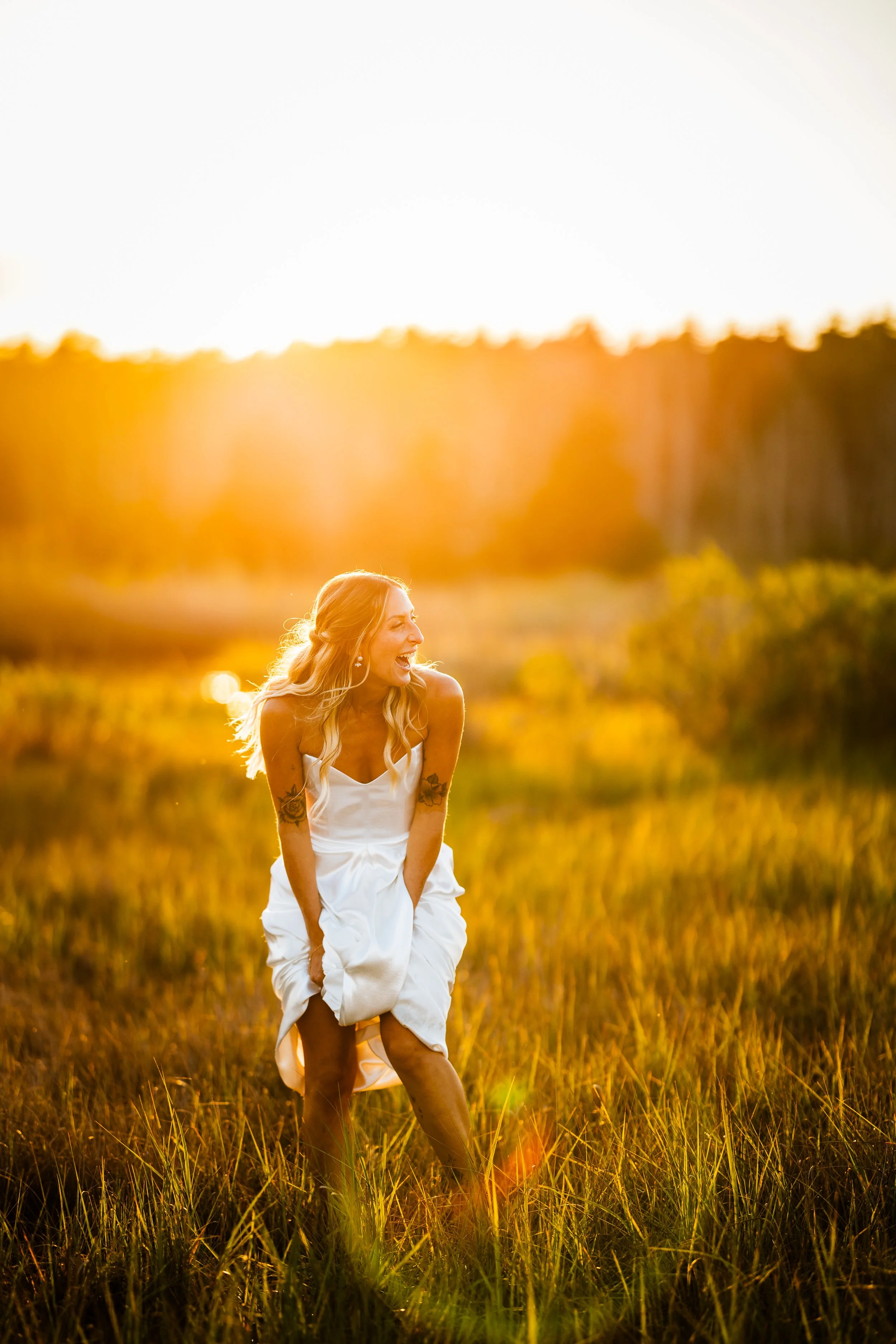 A woman in a white dress standing in a grassy field during sunset, laughing and holding up her dress.