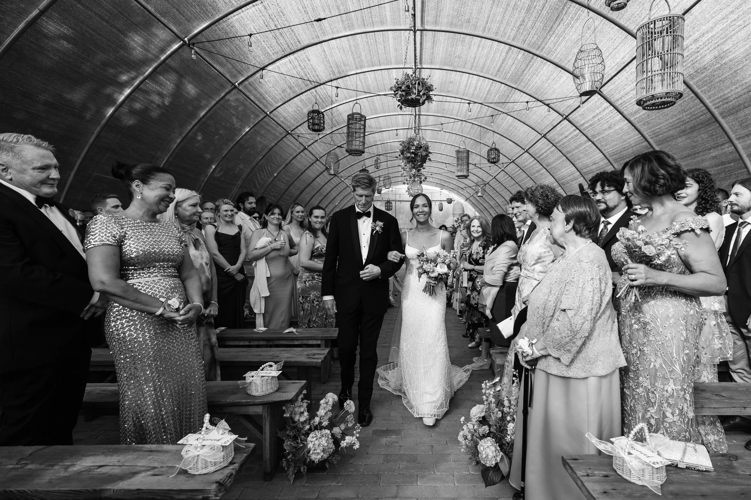 Black and white photo of a wedding ceremony with a bride and groom walking down the aisle inside a covered venue, surrounded by seated and standing guests.