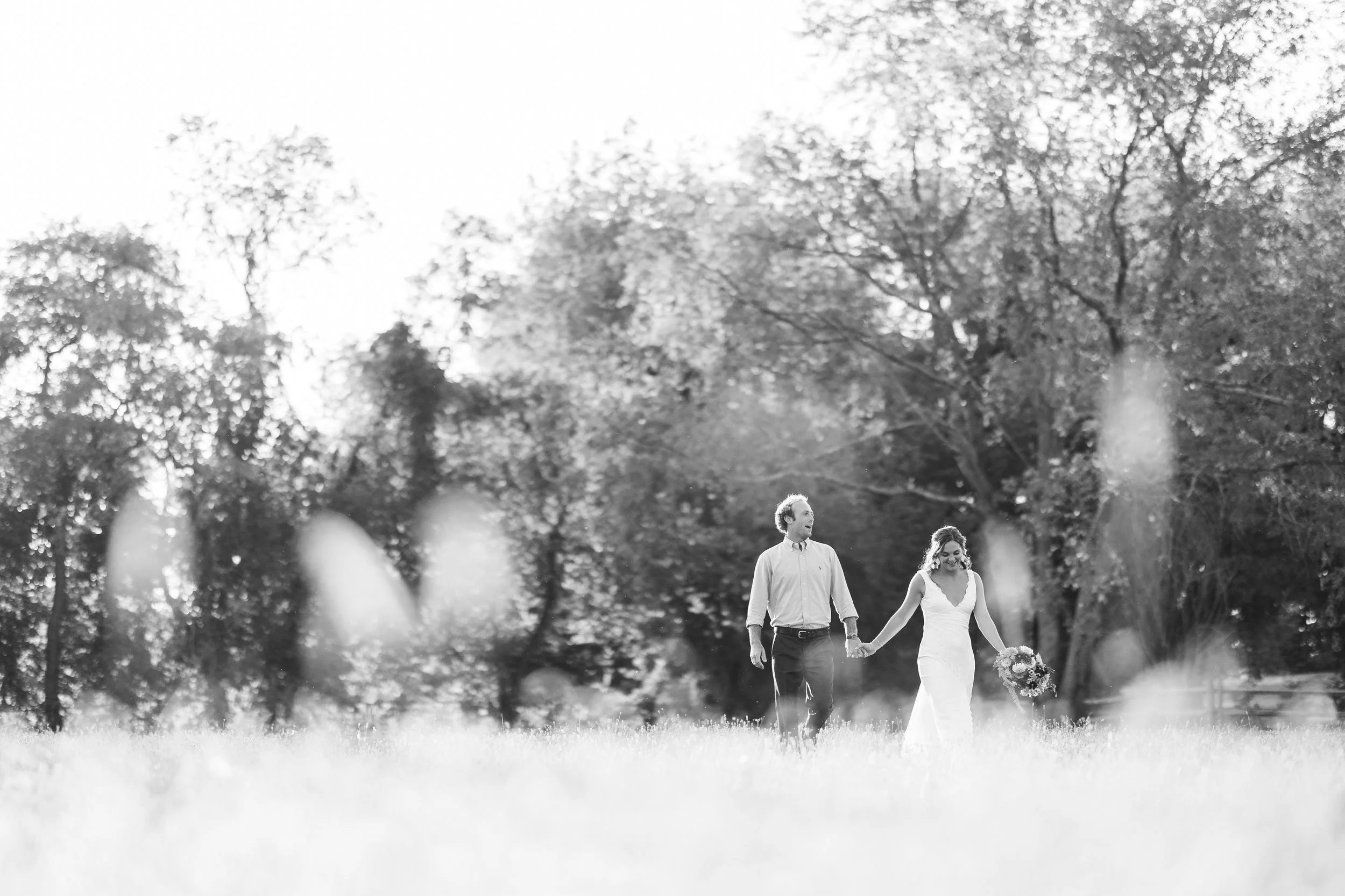 A black and white photo of a bride and groom holding hands and walking through a grassy field, surrounded by trees.
