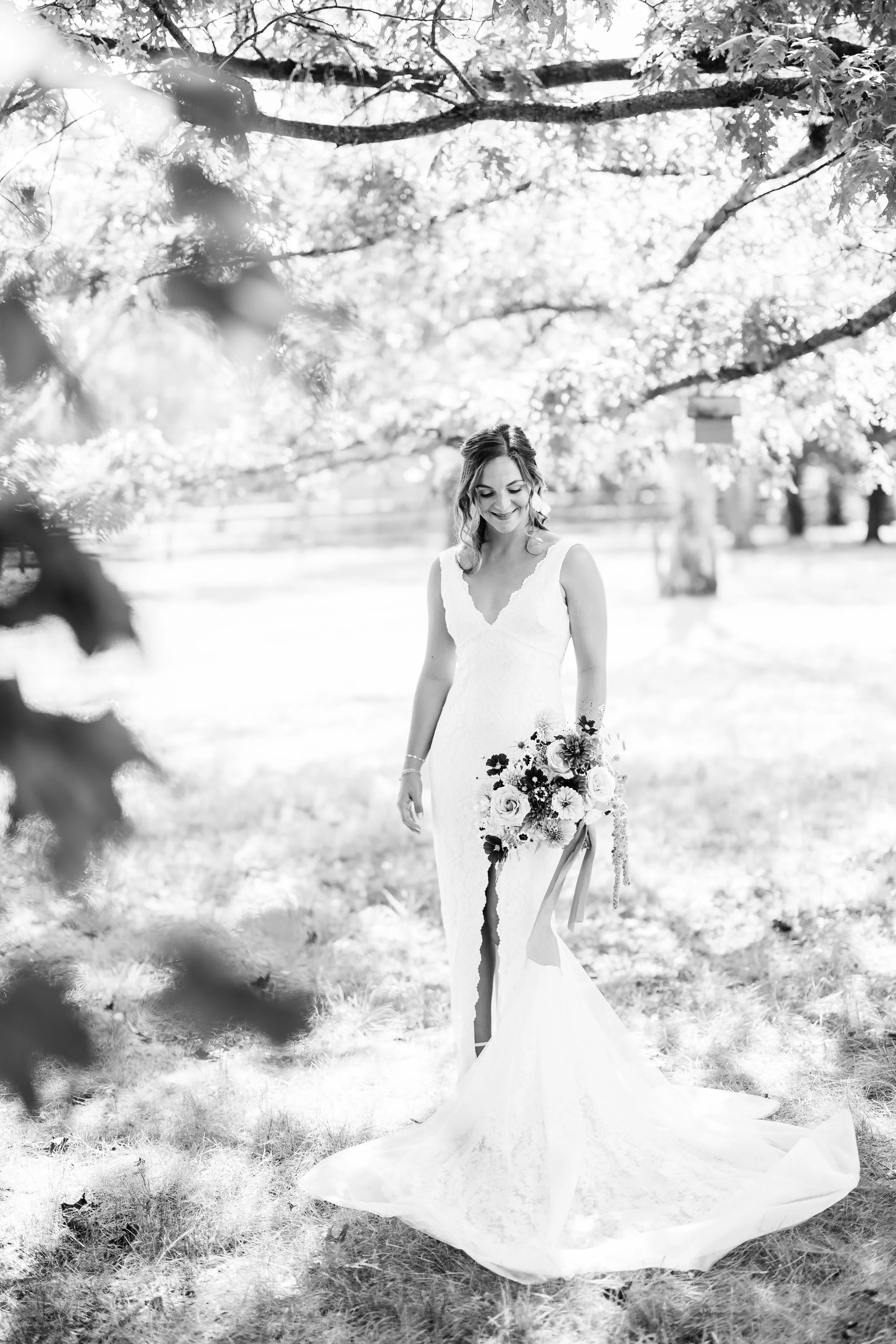 A bride in a sleeveless lace wedding gown standing outdoors under a tree, holding a large bouquet of flowers with a long ribbon, smiling while looking down.