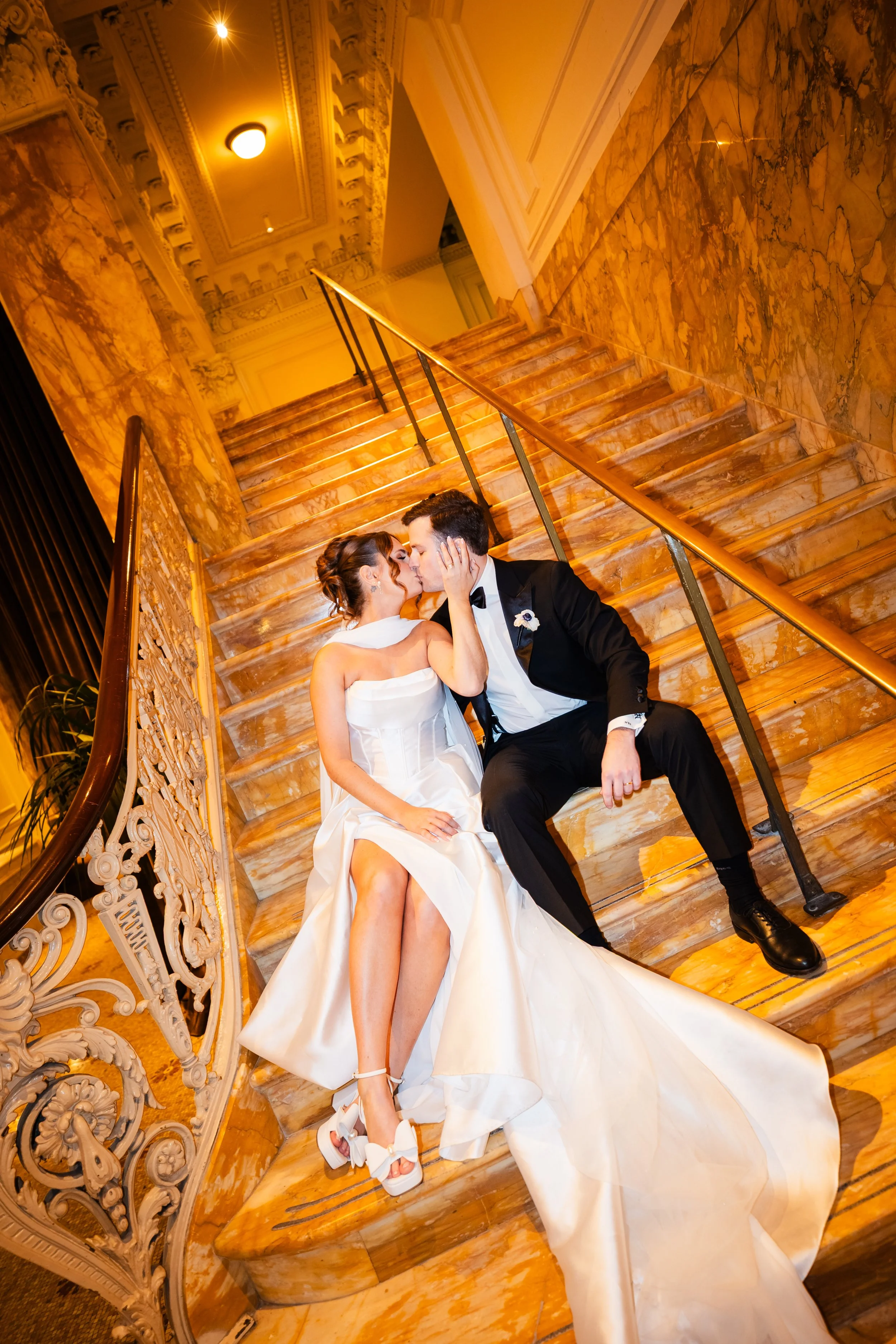 A bride in a white wedding gown and a groom in a black tuxedo kiss on a grand marble staircase with ornate railings inside an elegant building.