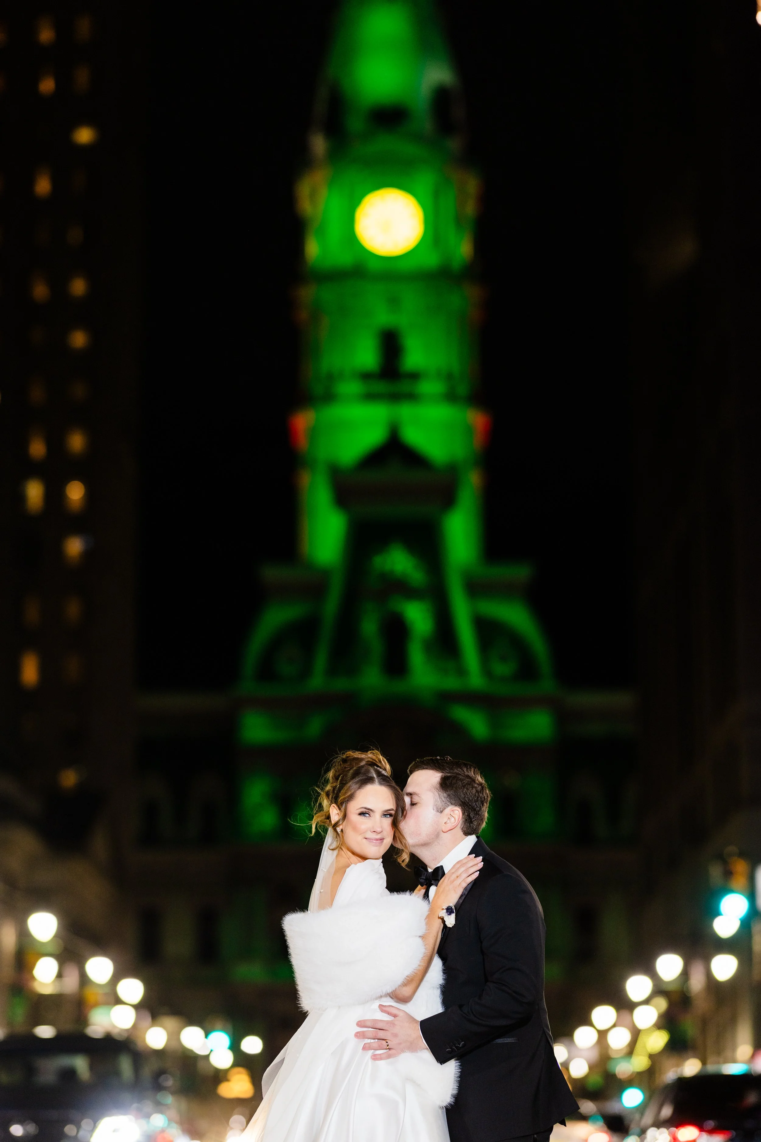 A bride and groom embrace at night in front of a tall, illuminated clock tower that is lit green.