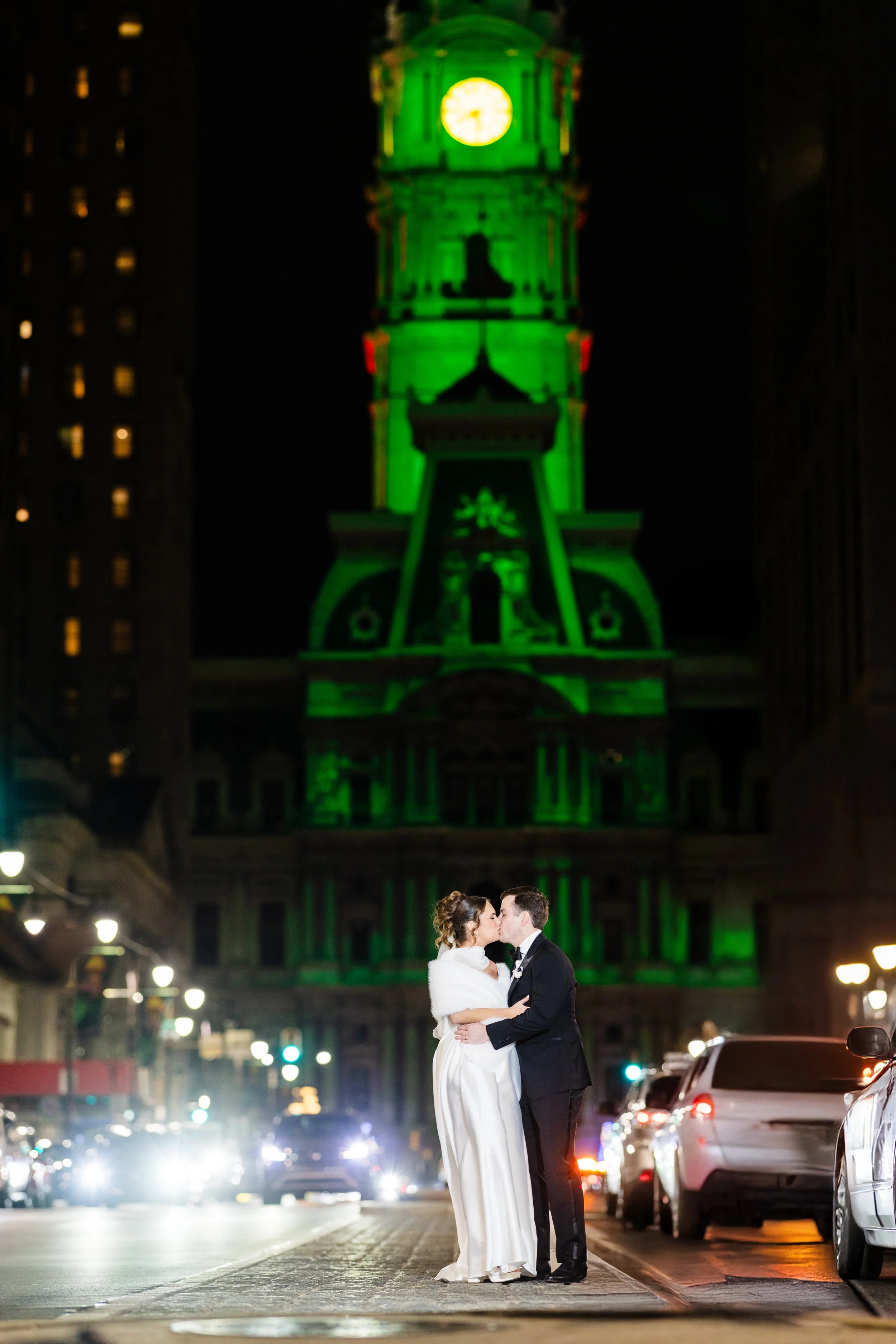 A couple in wedding attire sharing a kiss on a city street at night, with a large illuminated clock tower in green in the background.