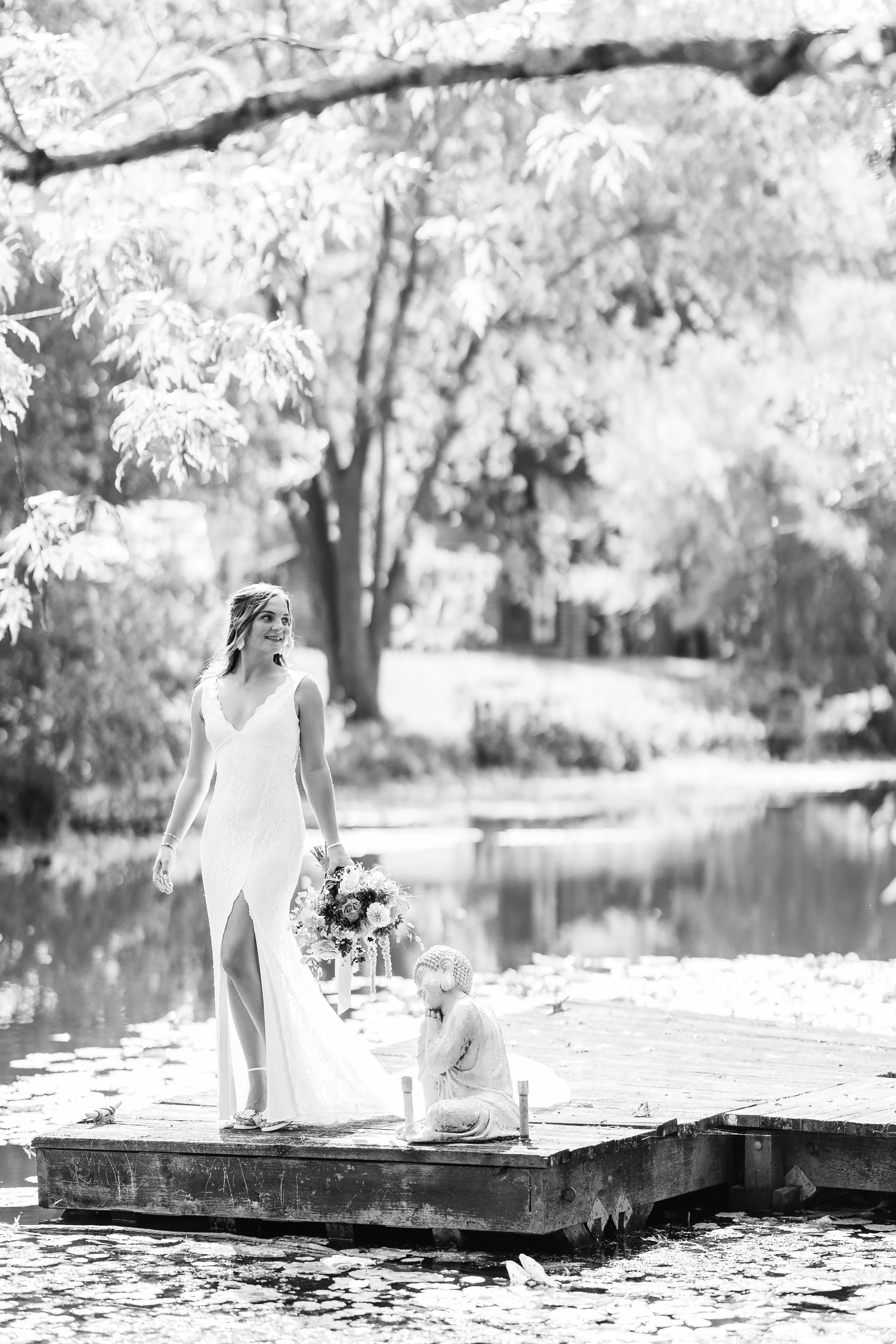 A woman in a white dress holding a bouquet walking on a wooden dock by a pond, with a sculpture of a kneeling child nearby, surrounded by trees and foliage in a park.