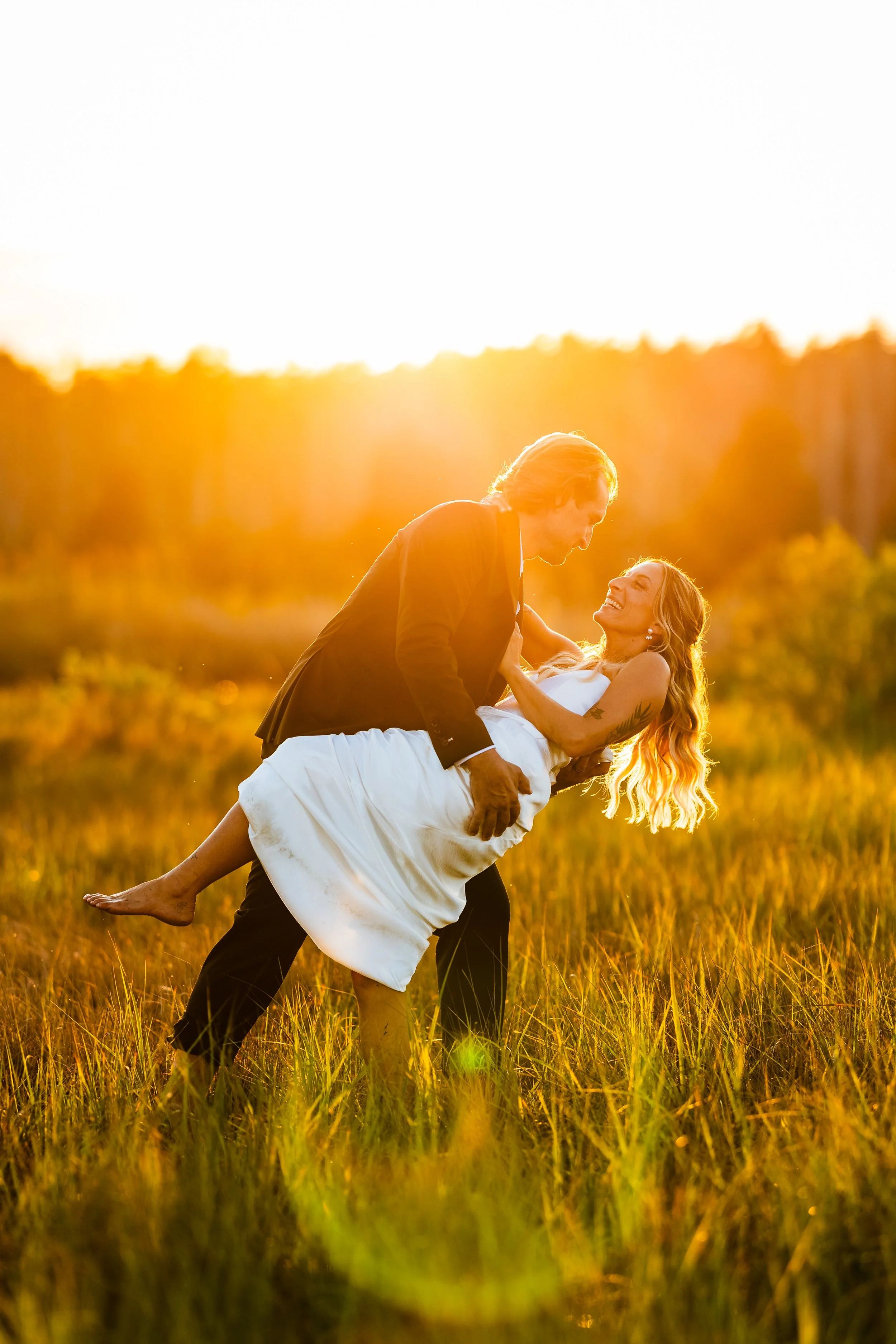 A man in a black suit holding a smiling woman in a white dress in a grassy field during sunset.