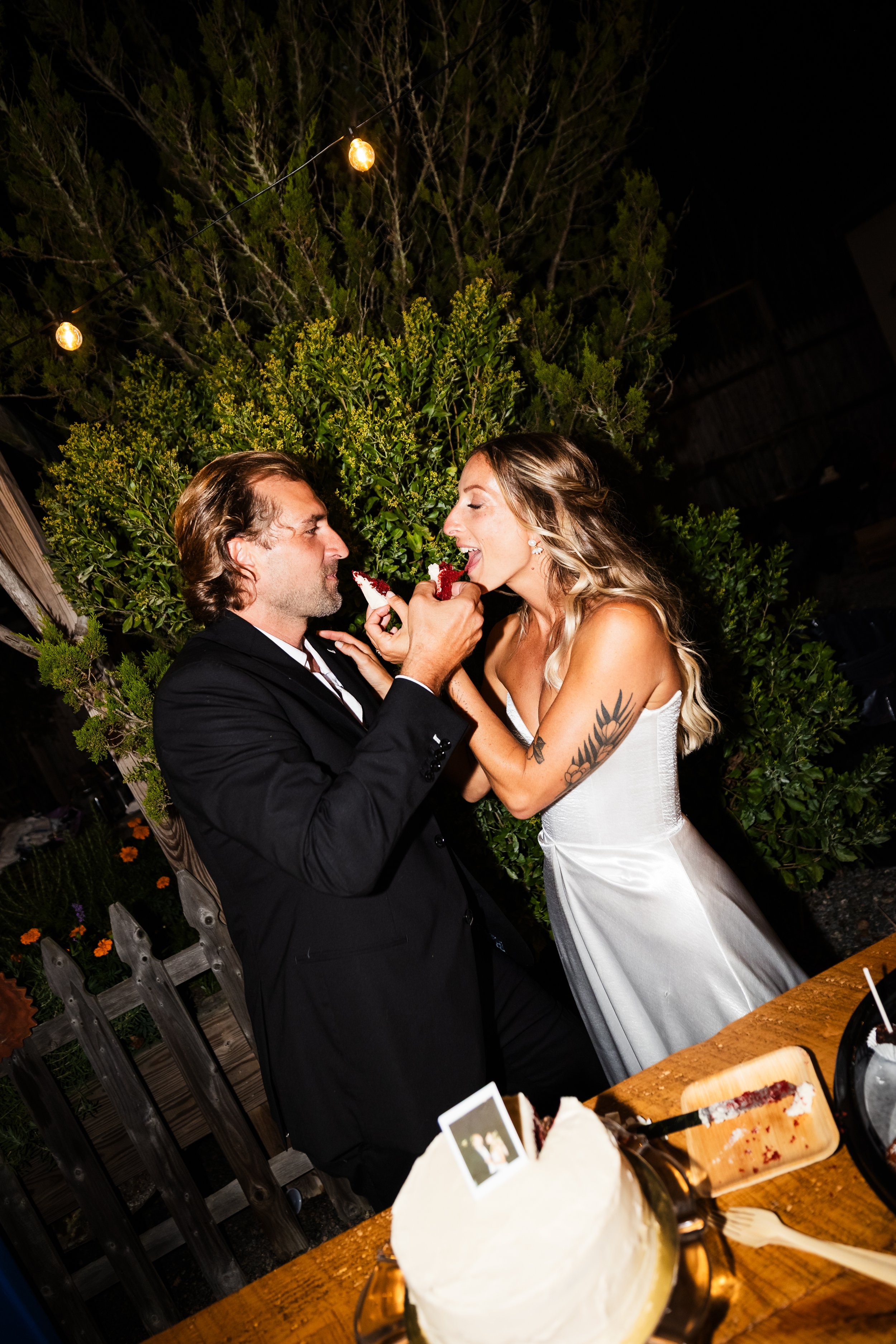 A wedding couple sharing cake outdoors at night with string lights and greenery in the background.