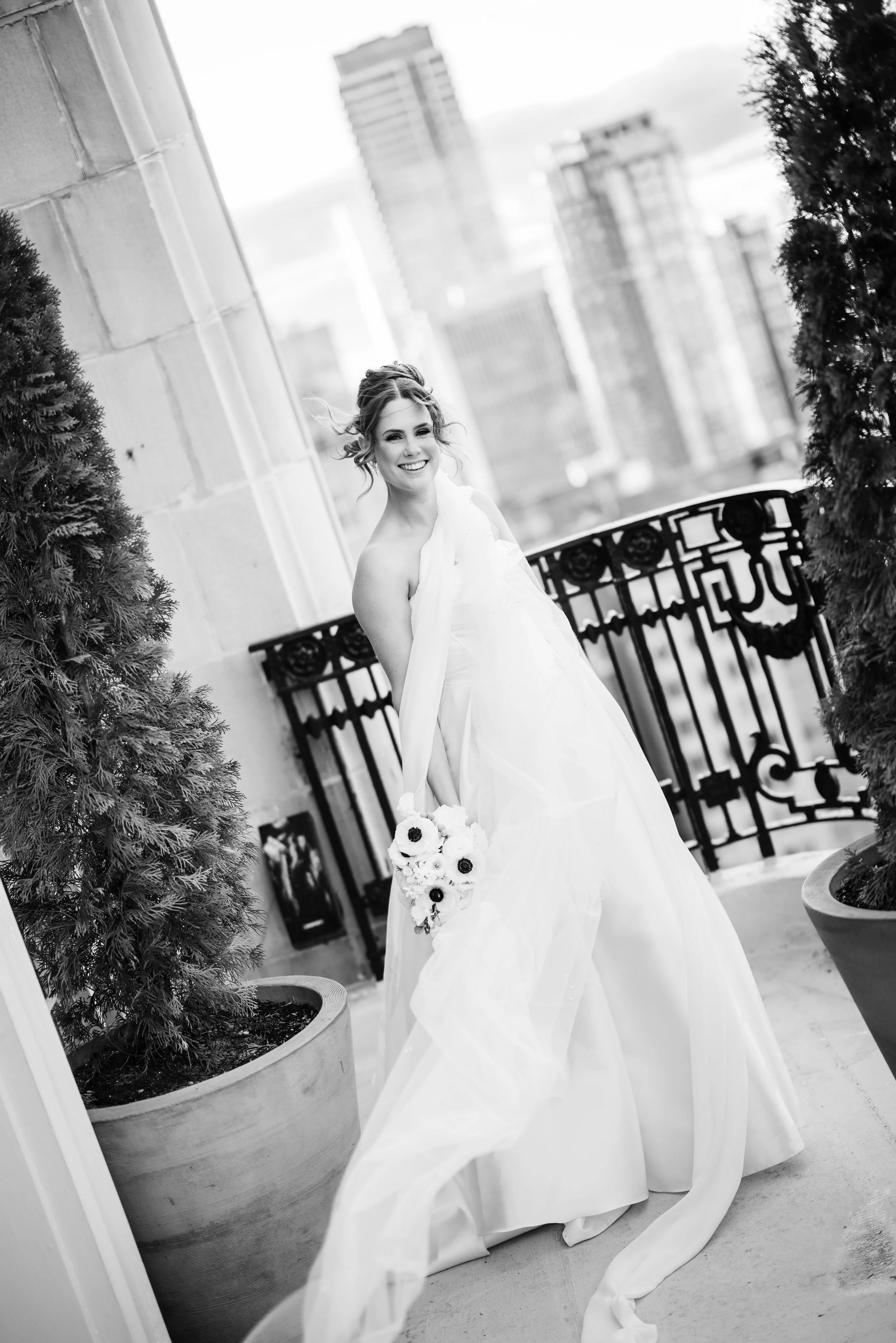 Black and white photo of a smiling bride in a wedding dress holding a bouquet, standing on a balcony with city skyline in the background.