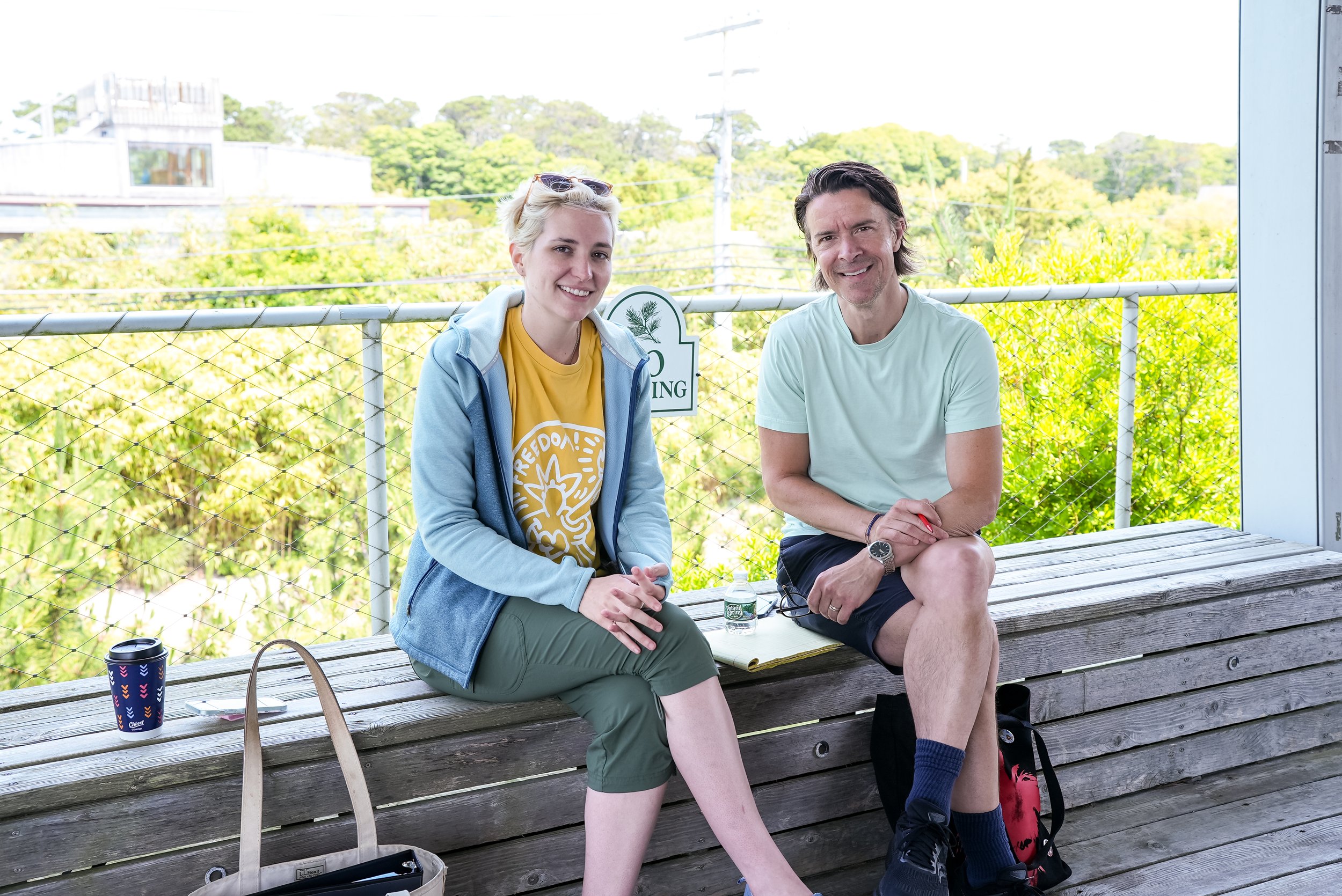 Two production staff members sitting cross legged against a fence with lush greenery behind them