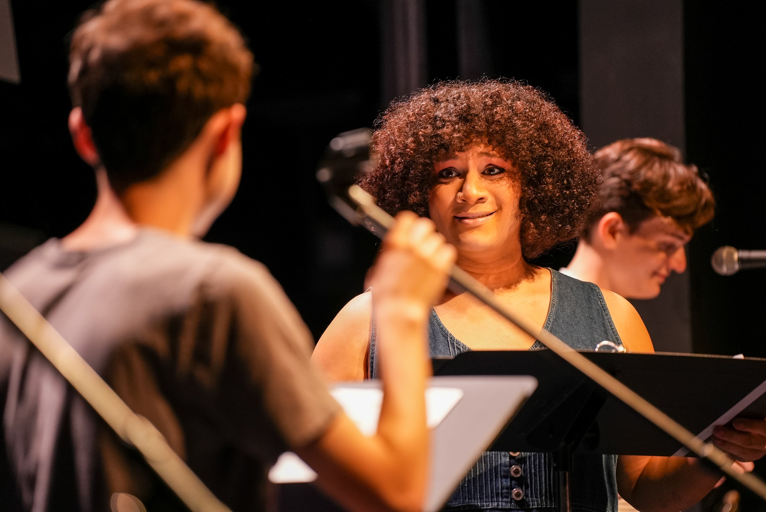 Three actors on stage with music stands and microphones conversing