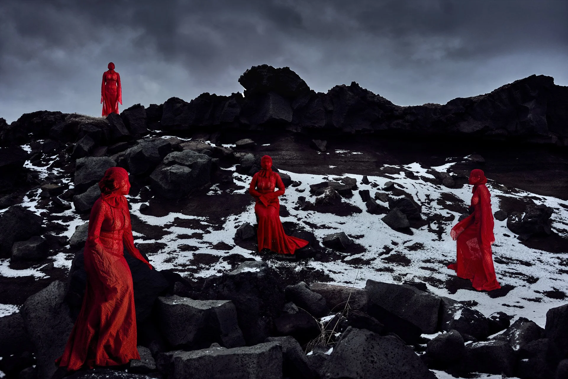 Four figures dressed in long red dresses and masks, standing and sitting on a rocky, snowy landscape under a dark, cloudy sky.