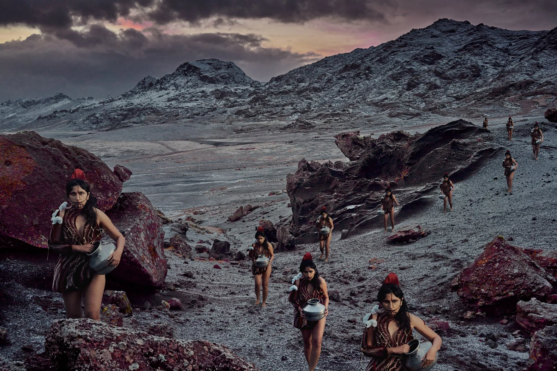 A surreal landscape with multiple women dressed in animal print outfits carrying bowls, scattered across a dark, rocky terrain with distant mountains and a cloudy sky.