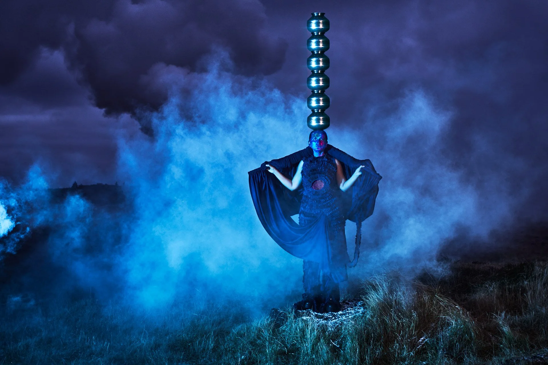 A person in dark clothing with body paint and a long black cape stands on grassy terrain at night, holding the cape open. Behind them, a tall, stacked metallic sculpture extends upward into stormy, dark clouds, illuminated with blue light, with swirling fog or smoke around.