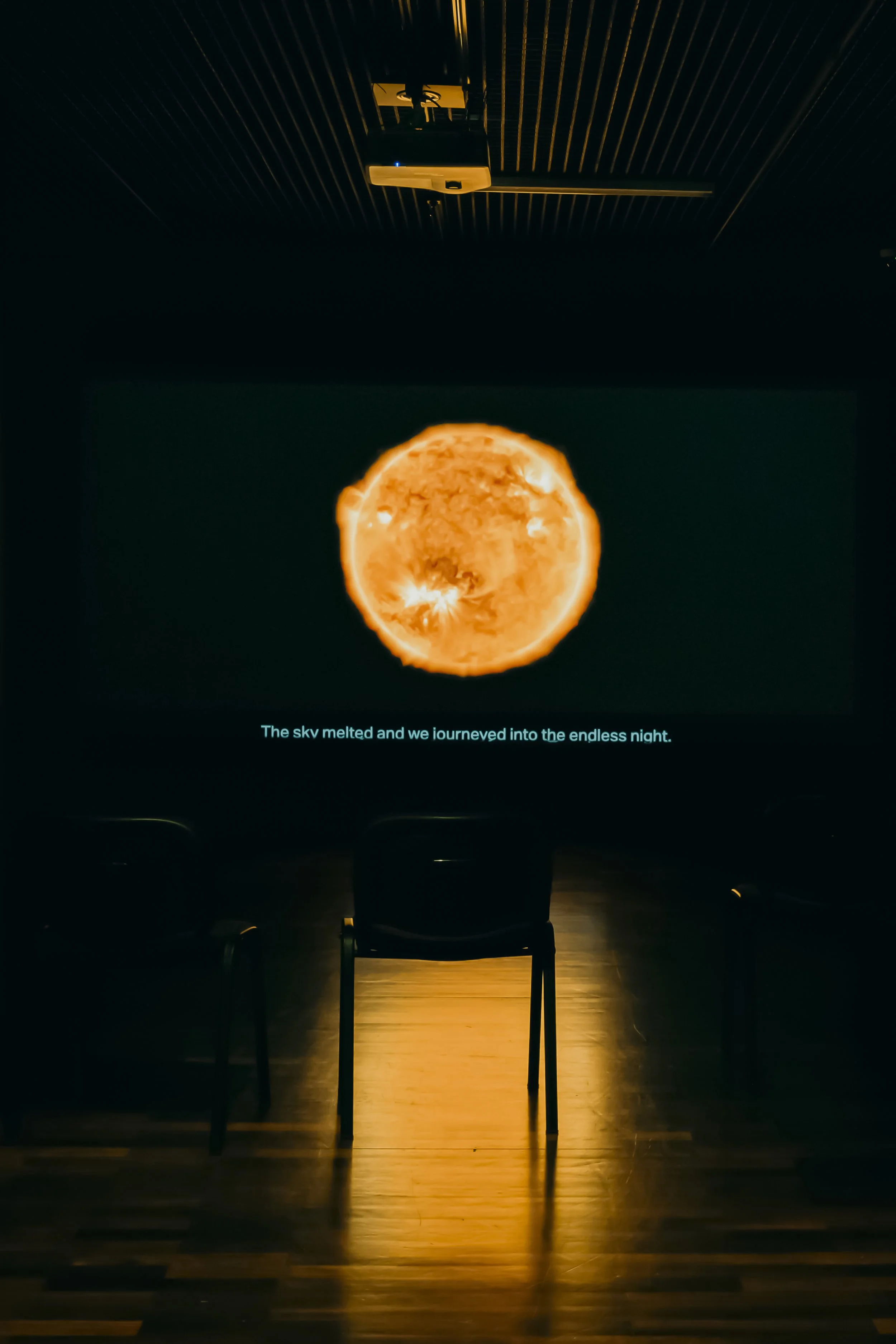 A dark room with a satellite projector displaying an image of the sun on a screen. The sun appears bright orange and glowing. There is a seat in the foreground, illuminated with a golden hue from the projection. Text at the bottom reads, 'The sky melted and we journeyed into the endless night.'