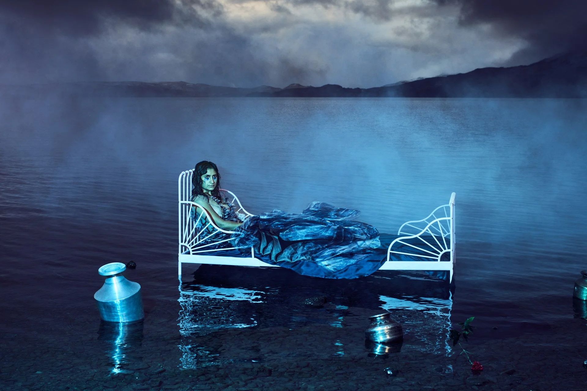 A woman lying on a white metal bed frame that is floating on water. The water is calm and reflects the bed. The woman has long dark hair, and is wrapped in a blue blanket. There are metal containers and a rose floating near the bed. The background shows a cloudy sky and distant mountains over a body of water.