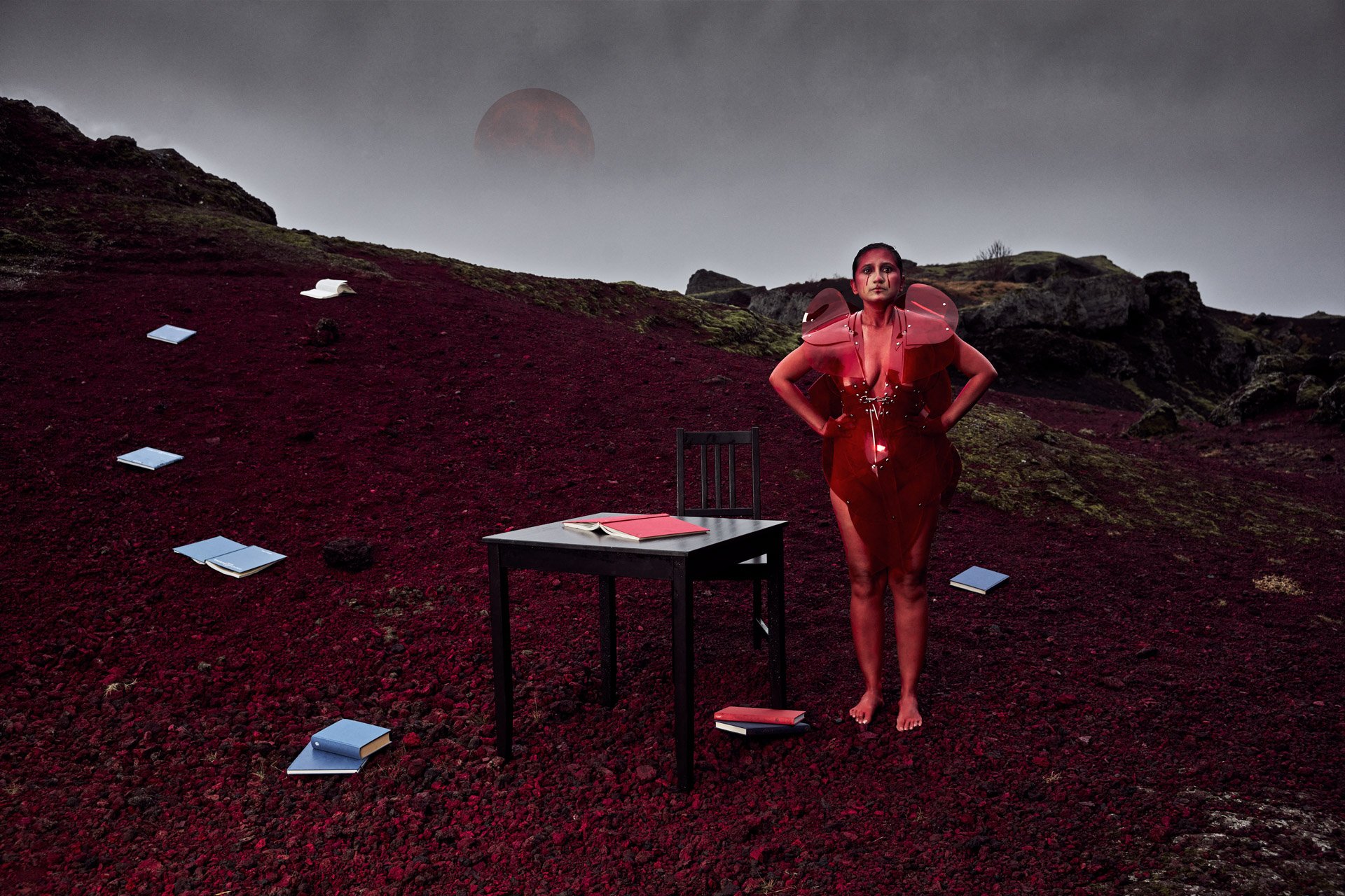 A woman in a red futuristic outfit standing barefoot on a red rocky landscape with scattered books and a black table, under a dark sky with a large red moon or planet in the background.