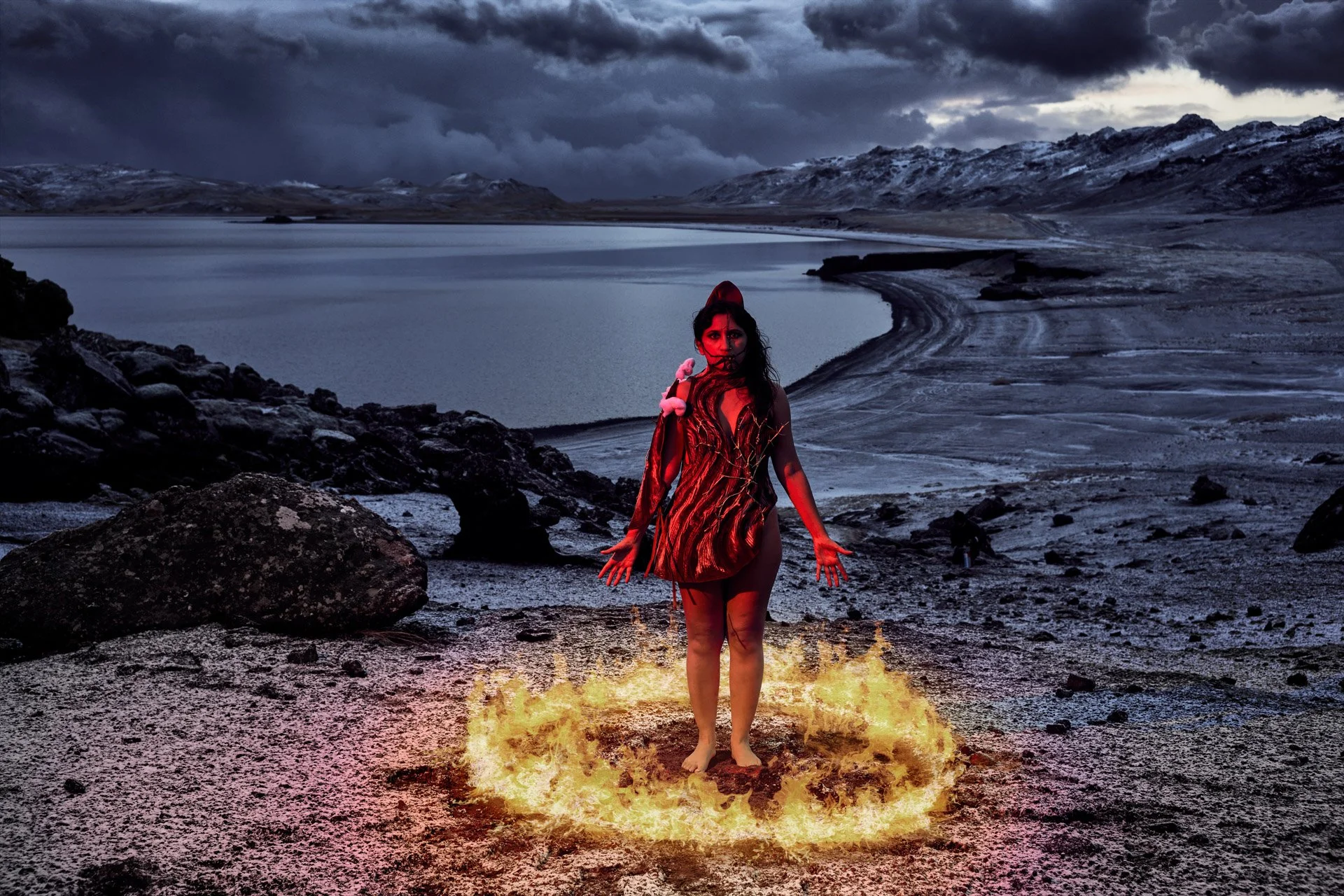A woman in a red dress stands barefoot on a patch of fire on a rocky shoreline, with a lake and snow-capped mountains under a dark, cloudy sky in the background.