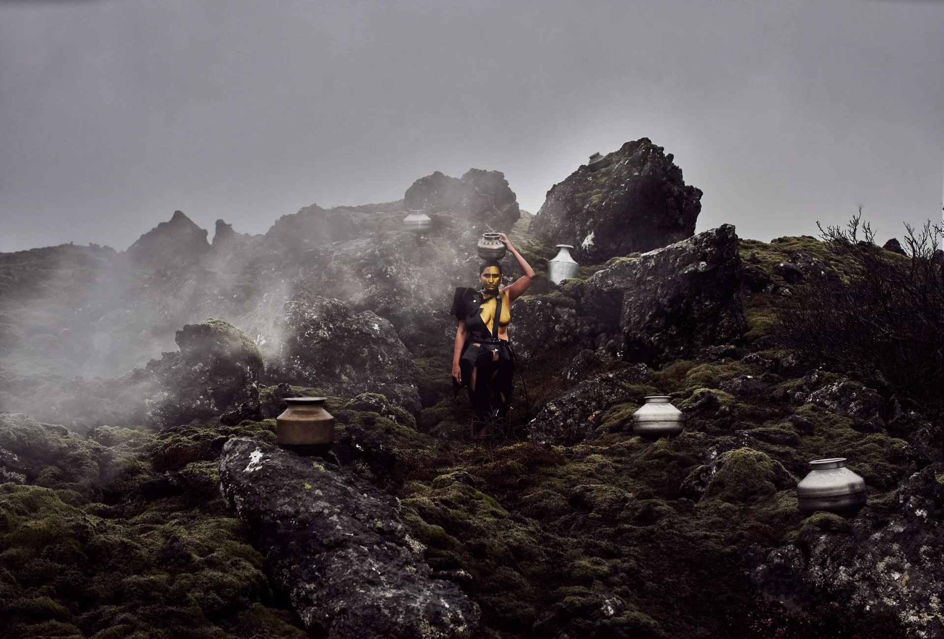 A woman in a black and yellow outfit carrying pots on her head walking across a rocky, moss-covered landscape with mist and gray sky.
