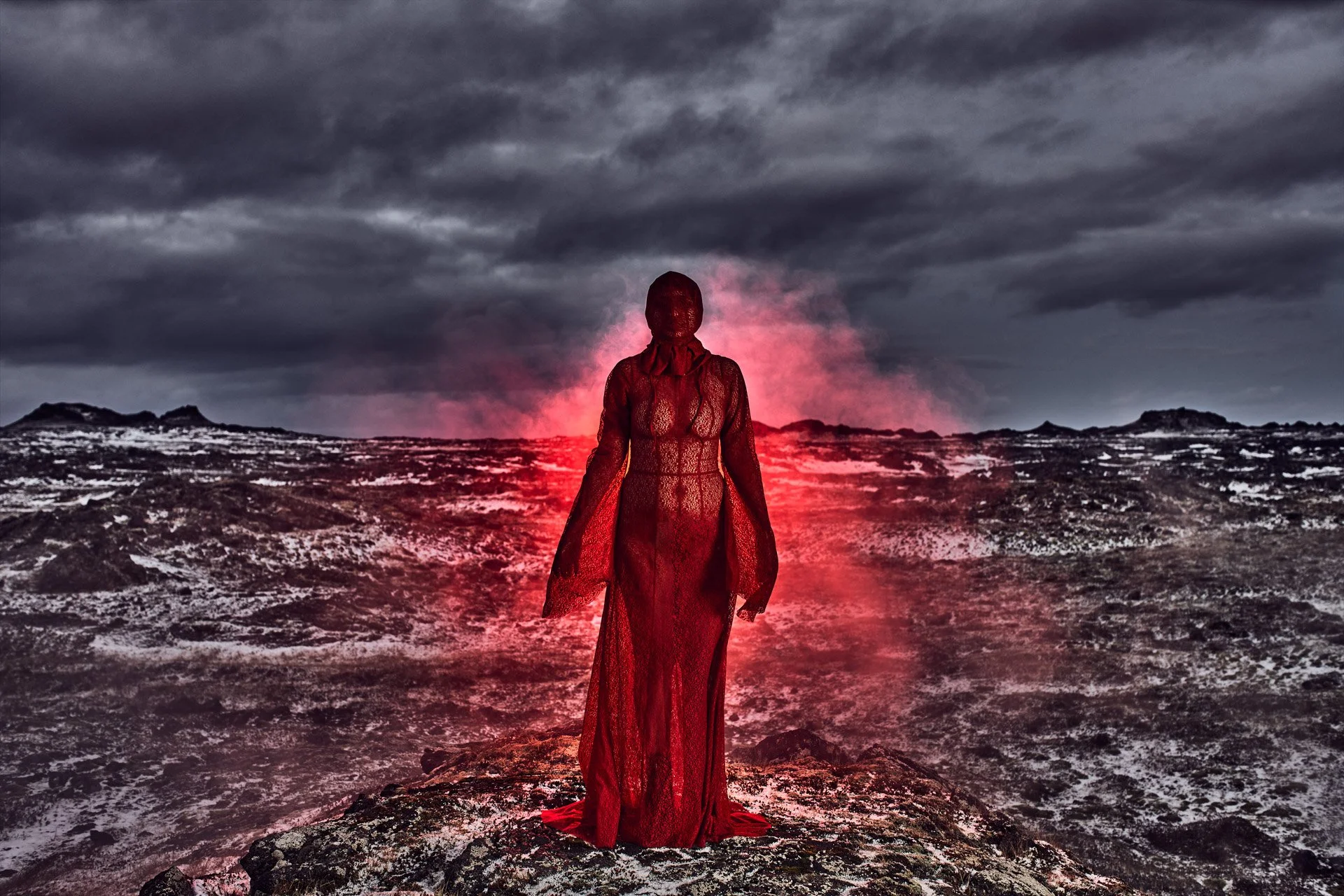 A woman in a red lace dress standing on a rocky terrain, facing away, with a glowing red light or smoke behind her under a dark cloudy sky.
