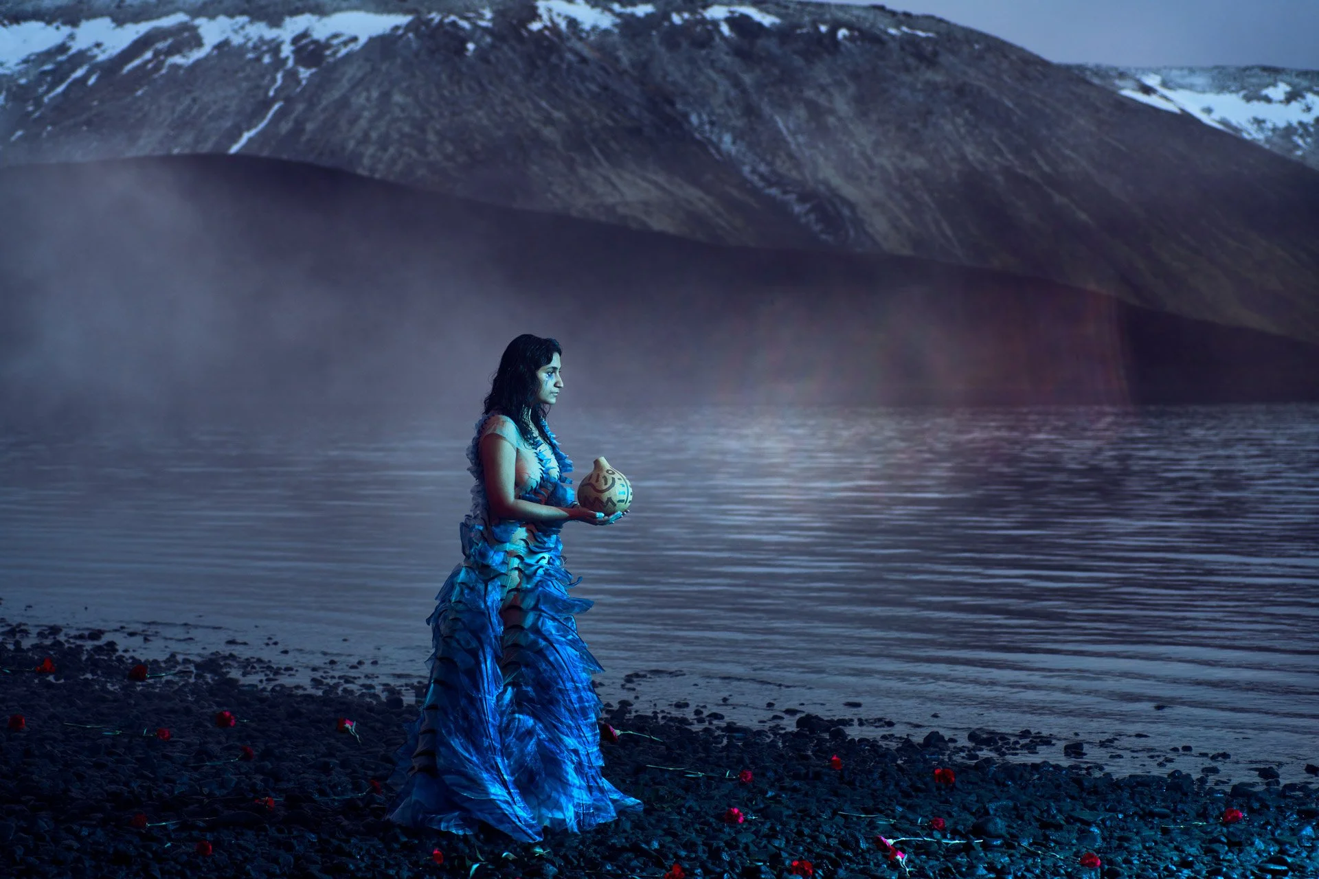 A woman in a flowing blue dress holding a decorative vessel, standing by a calm body of water with a mountainous backdrop, with red flowers scattered on the rocky shore.