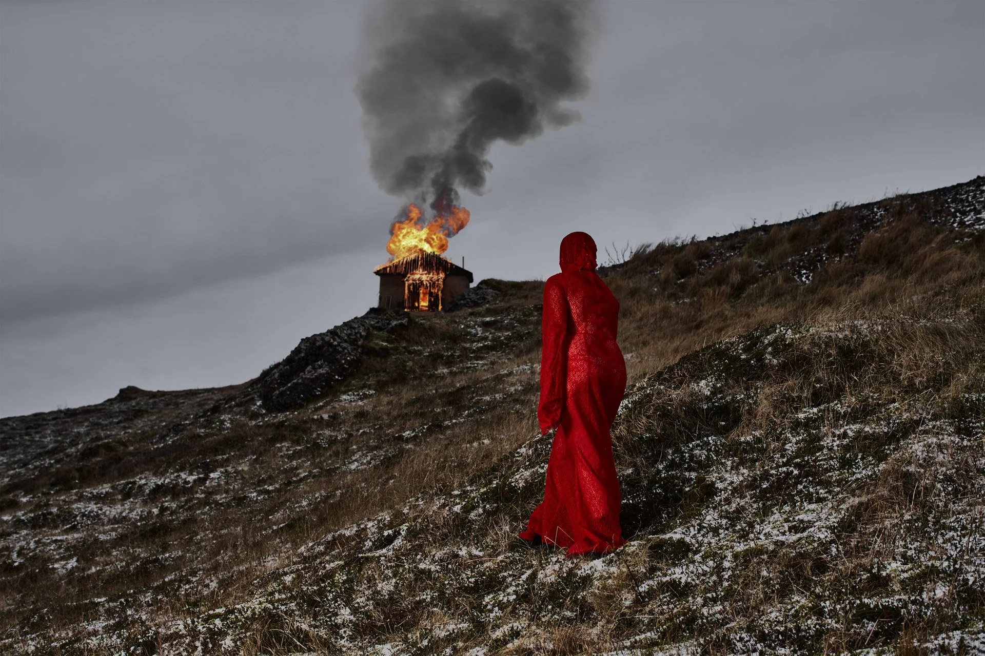 A woman dressed in a long red gown standing on a snowy hillside, watching a house on fire in the distance.