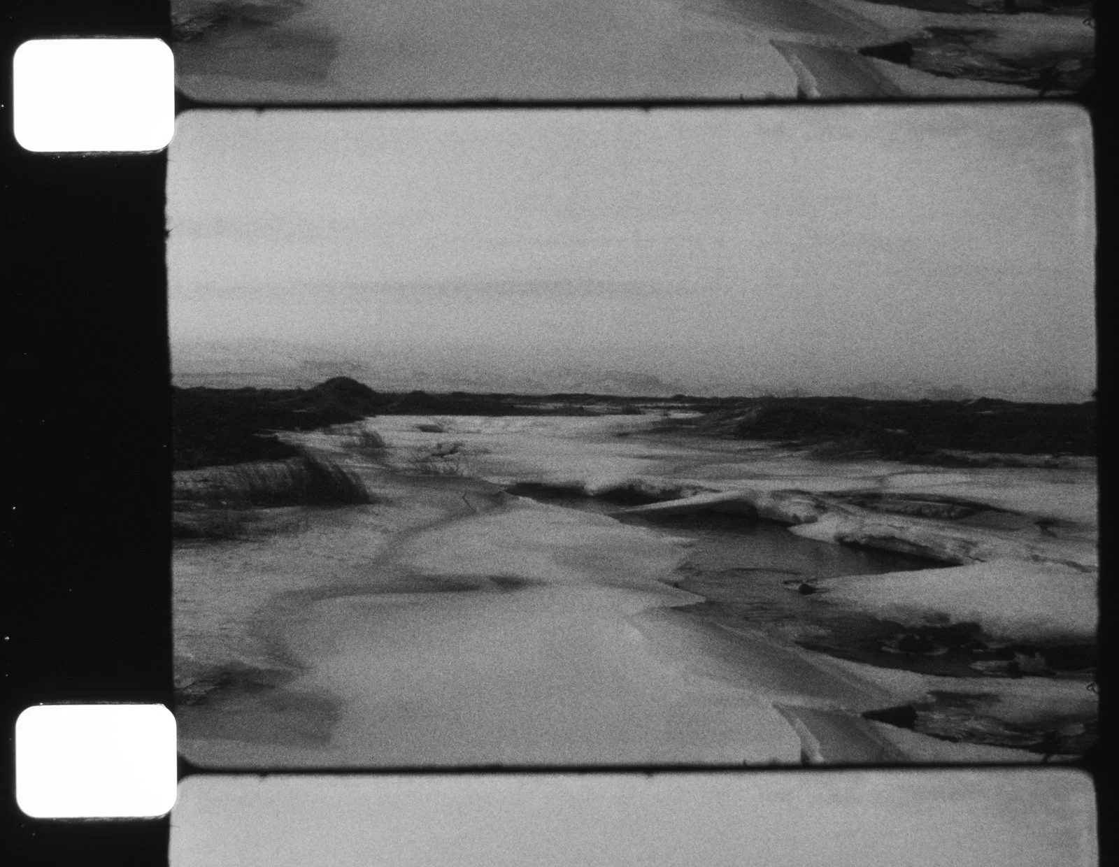 Black and white aerial photograph of an arid, rocky landscape with some water bodies and distant hills or mountains under a cloudy sky.