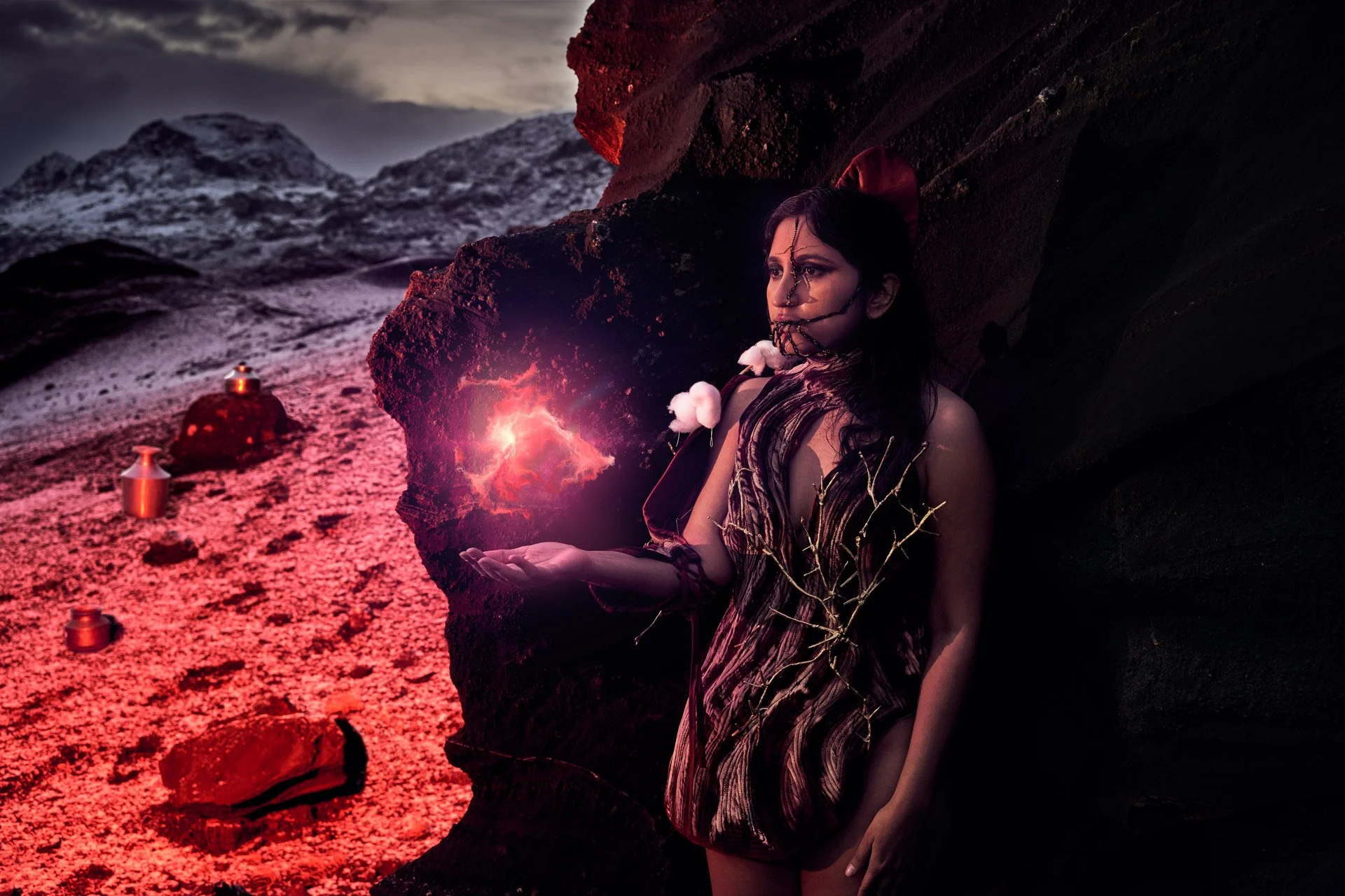A woman in a patterned dress with a dark landscape background, holding a glowing, lightning-like object in her hand, with candles around her and mountains in the distance at sunset.